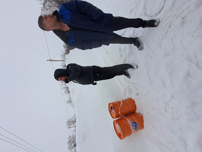 The image depicts an outdoor, snow-covered scene in the city of Полянки, Russia, under an overcast sky. Two adult males are standing on a snowy path or road. To the left, a man wearing a dark hooded puffer jacket, dark pants, and gloves has his right hand raised towards his mouth. To his right and slightly forward, another man wears a blue and dark blue winter jacket with a dark fur collar, dark pants, and dark footwear; he is looking off to the right, and a gold-colored necklace is visible around his neck. Two orange cylindrical barrels, partially covered in snow and wrapped in clear plastic, rest on their sides to the left of the first man; one barrel displays a blue and white label. The ground is thickly covered in snow, with visible footprints around the men. A circular manhole cover is partially visible in the snow near the second man's feet. In the background, large snowdrifts rise, behind which bare trees and utility poles with power lines are visible.