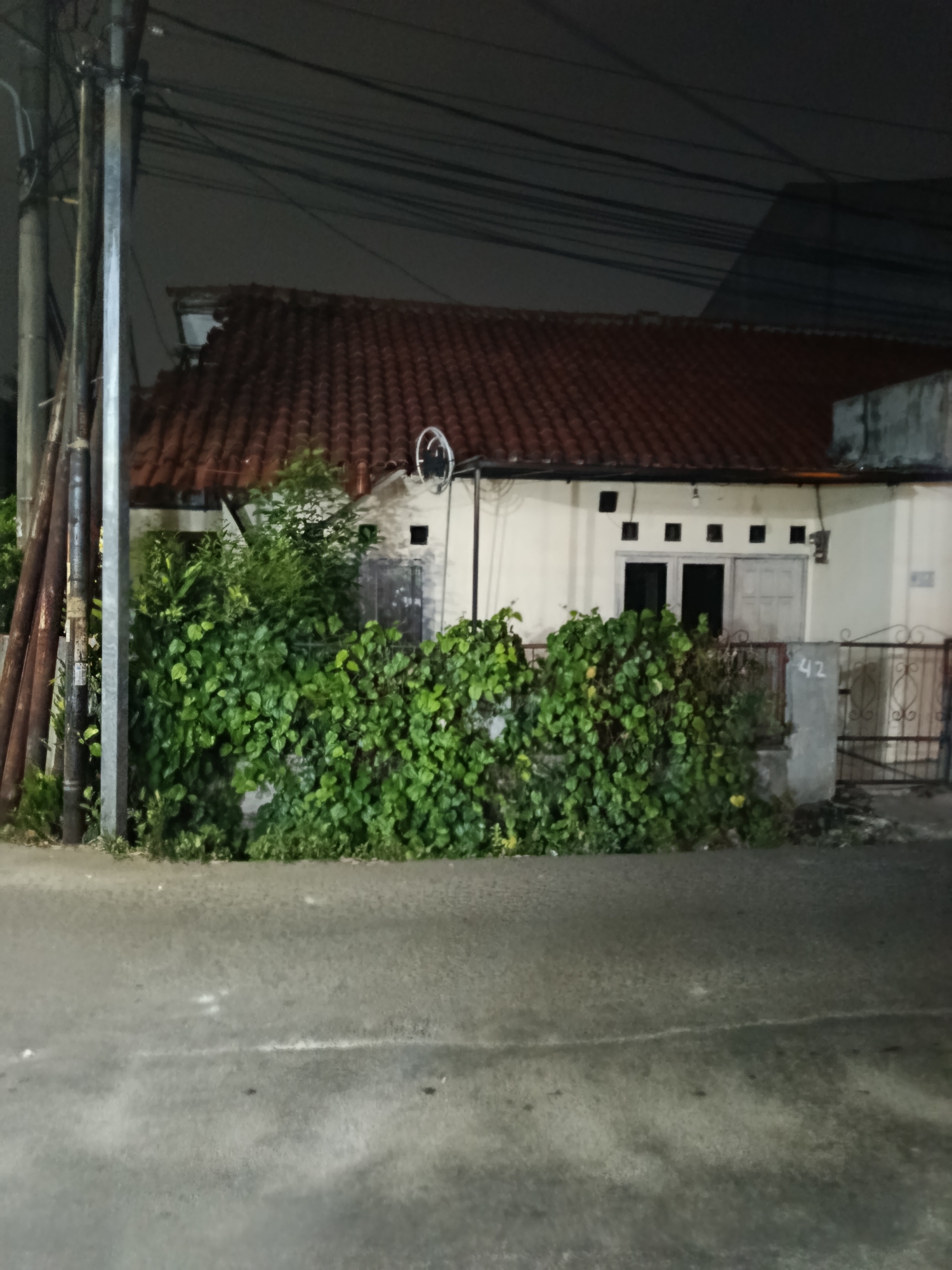 The image depicts a residential house in Depok, Indonesia, at night. The house has a terracotta tile roof and a light-colored facade with several small, square windows. A dense green hedge covers the front of the property, obscuring much of the house. To the left, there are several vertical poles, possibly part of a utility structure, and overhead power lines are visible against the dark sky. A metal fence with an ornate design is present in front of the house, and a grey pillar with the number "42" painted on it stands next to the fence. The foreground shows a wet asphalt road, suggesting recent rain. No people or ongoing actions are visible in the scene.
