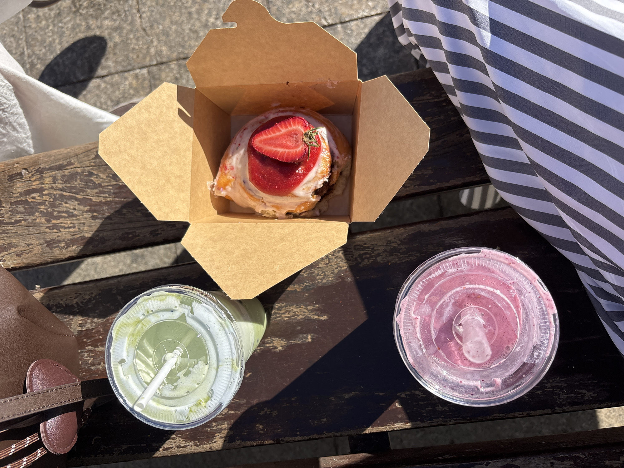 On a weathered wooden surface, likely a bench, are two beverages in clear plastic cups with straws and a cardboard box containing a pastry topped with strawberry. The drink on the left appears green, potentially matcha-flavored, while the drink on the right is pink. Adjacent to the green drink is a brown handbag. To the right of the box is a white and gray striped cloth. Based on the warm sunlight and the food and drink items, the scene is likely set outdoors. While the exact location within Madrid cannot be determined from the image alone, the presence of outdoor seating and a pastry could suggest a café or park.