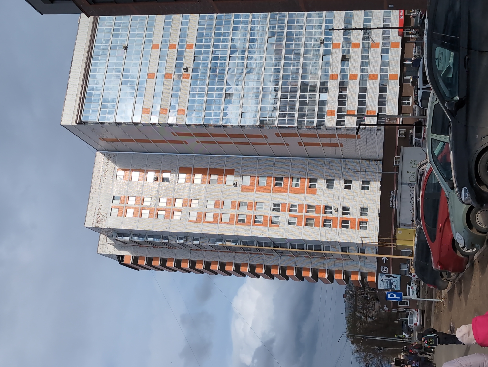This image depicts an urban street scene with several multi-story buildings under an overcast sky. The most prominent structure is a tall, modern building with a facade primarily made of reflective glass panels, which mirror the cloudy sky. Below it, another multi-story building with a lighter, textured exterior features rows of smaller windows interspersed with orange or brownish rectangular accents. A third, shorter building is partially visible in the background, behind parked cars.
In the foreground and to the right, a street or parking area is populated with several parked vehicles, including a dark grey car, a red car, and a light-colored SUV. Multiple people are visible walking in the lower right, some wearing winter clothing, such as coats and a pink hat, indicating cool weather.
Notable details include air conditioning units on the building exteriors. Street signs are present; one sign, attached to a pole, reads "ОБИ СПРОДАЖ НЕДВИЖИМОСТИ" (OBI Sales of Real Estate) and another displays "МАГНИТ" (Magnit), a Russian supermarket chain. A blue square sign with a white 'P' designates parking. The time of day appears to be daytime, though the heavy cloud cover prevents direct sunlight. The scene suggests an everyday urban environment, likely in a Russian-speaking region given the Cyrillic text.