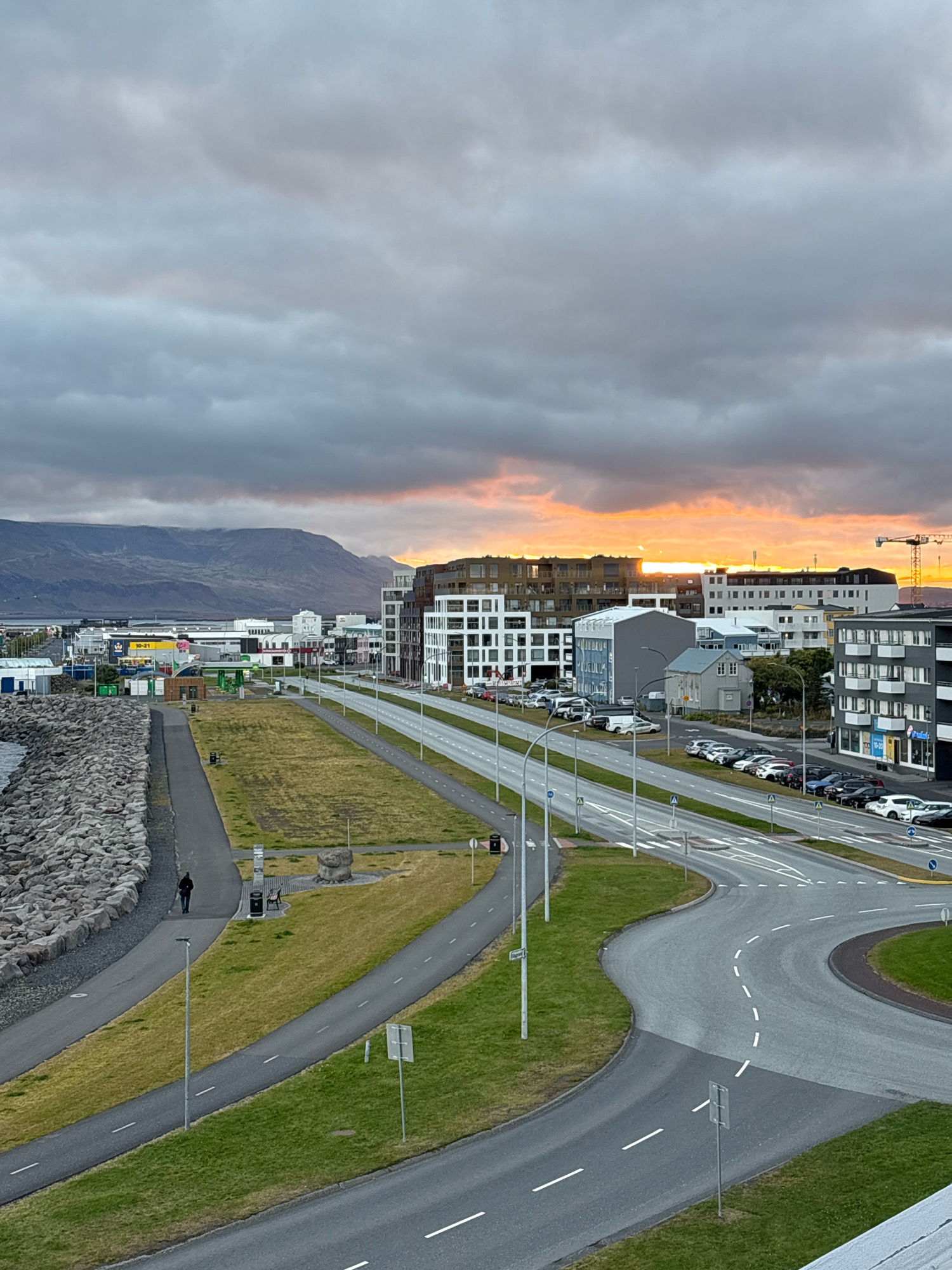 The image depicts a cityscape in Reykjavik, Iceland, likely during sunset or sunrise, given the warm glow on the horizon and the cloudy sky. In the foreground, a man is walking on a paved pathway beside a rocky shoreline. Beyond the pathway, a grassy area with a bench and a solitary figure is visible. A road with multiple lanes curves through the scene, with parked cars lining one side. In the background, modern buildings with a mix of glass and concrete facades dominate the skyline. Further in the distance, a mountain range is silhouetted against the sky. The overall impression is one of urban development meeting natural landscapes under a dramatic sky.