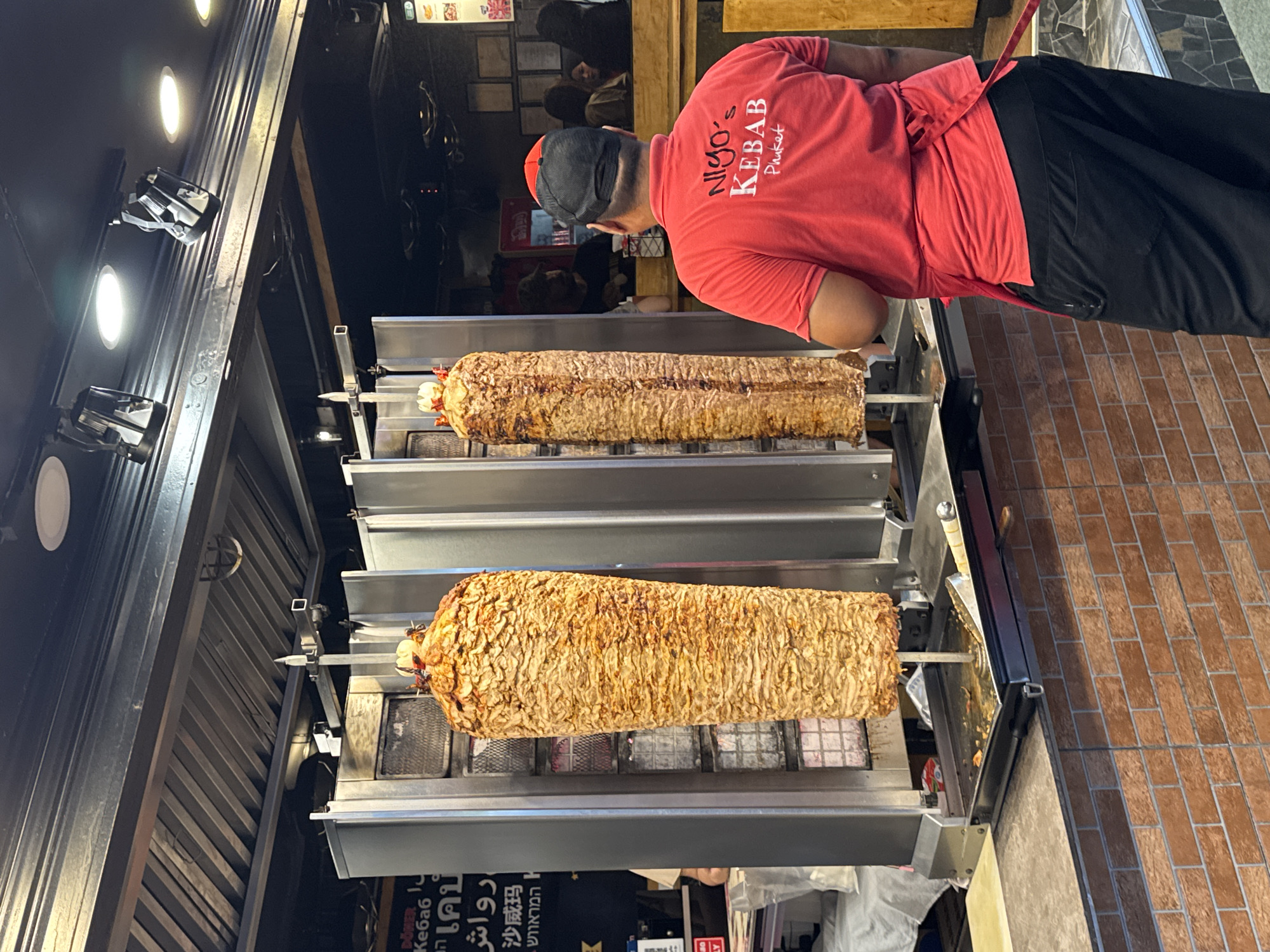 In Patong, Thailand, a man wearing a red t-shirt with the logo "Niygo's Kebab Phuket" and a black baseball cap stands in front of two vertical rotisseries. Each rotisserie holds a large, cylindrical stack of thinly sliced, cooked meat, characteristic of döner kebab. The man is facing the rotisseries, with his back to the camera, suggesting he is either preparing to serve or is a staff member. Behind him, other people can be seen in the background of the establishment, which appears to be a food stall or restaurant. The environment is lit by overhead lights, and the scene is rich with the visual details of a busy food service area.