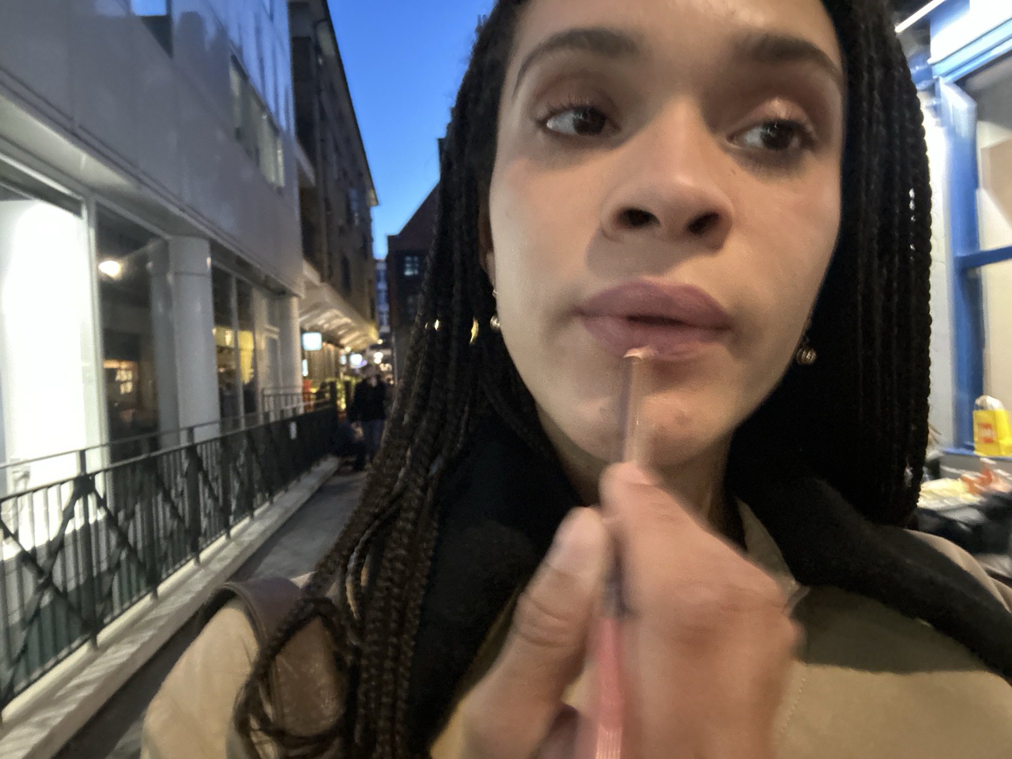 A woman with dark braided hair is applying lip liner in an outdoor urban setting. She is fair-skinned and has small gold earrings. The background shows a London street at dusk, with buildings on either side. On the left is a modern building with large windows. A metal railing runs alongside the sidewalk. In the distance, people are walking along the street. The lighting is a mix of natural and artificial from streetlights and building interiors. The woman appears to be standing on the sidewalk as she applies her makeup.