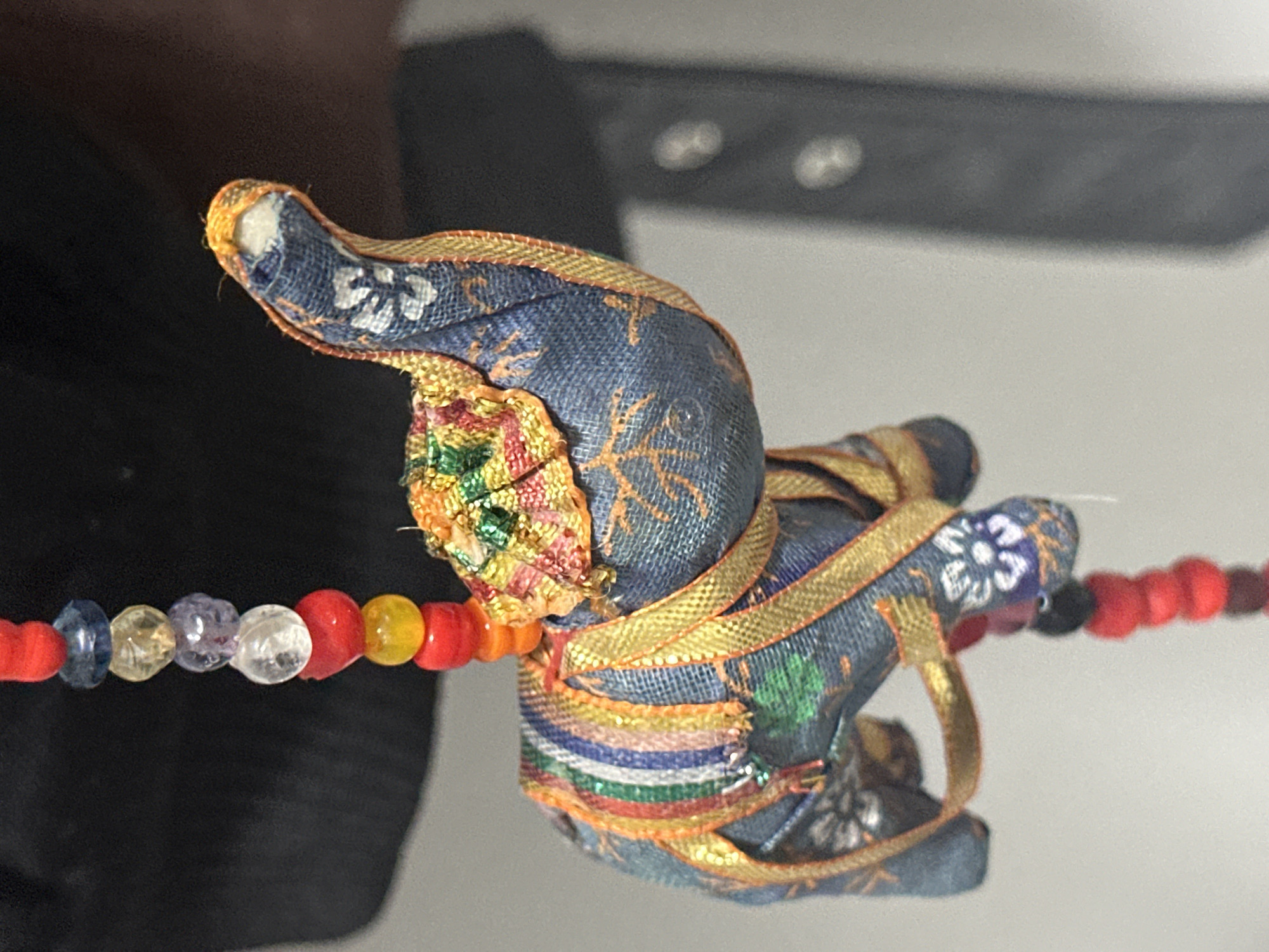 The image shows a close-up of a decorative fabric elephant ornament. The elephant is blue with orange, gold, and multi-colored striped embellishments. It is hanging from a strand of colorful beads, including red, yellow, orange, clear, purple, and blue beads. The background is out of focus and appears to be a dark fabric object and a light gray surface. There is no indication of the city of Nevinnomyssk, Russia, or any other specific location.