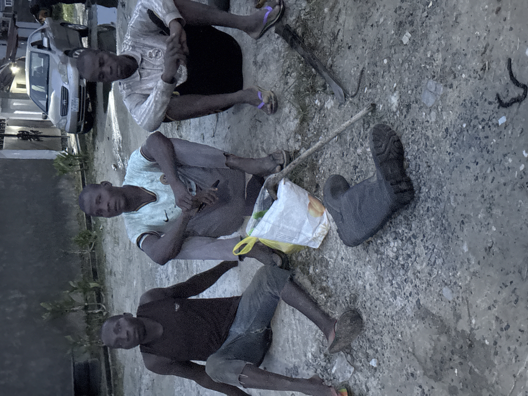 Three adult males are depicted sitting on the ground outdoors, appearing to be resting or observing. The image is rotated 90 degrees counter-clockwise.
The man in the top portion of the frame (when viewing the image in its current orientation) wears a white long-sleeved shirt with a "REAL MADRID" logo, dark shorts, and purple flip-flops. His hands are clasped in front of him. The man in the middle wears a light-colored t-shirt with a "Nike" logo and dark trousers. He holds a mobile phone in his right hand. The man in the bottom section wears a dark t-shirt and dark, possibly distressed, shorts or cropped trousers. He is barefoot. All three individuals appear to be looking forward, possibly in the same direction, though their expressions are neutral.
They are seated on uneven, rocky, and possibly muddy or dusty ground. Several objects are scattered around them, including a long wooden stick or pole, a single grey work boot or rubber boot, and a white plastic bag with yellow handles, which appears to contain groceries like bread. To the right of the middle man, what looks like a machete or similar tool with a wooden handle is visible on the ground. Black, coiled debris is also present on the ground further to the right.
In the background, a silver-colored sedan, resembling a Toyota Corolla, is parked on what appears to be a paved or unpaved road. A partially visible Nigerian license plate on the car shows "ABJ", which typically indicates Abuja, though the provided context is Iranla, Nigeria. Some small green plants and building structures can be seen in the background, suggesting a semi-urban or populated setting. The lighting suggests it is daytime, possibly overcast or late afternoon. No specific activity beyond resting or waiting is clearly depicted.
