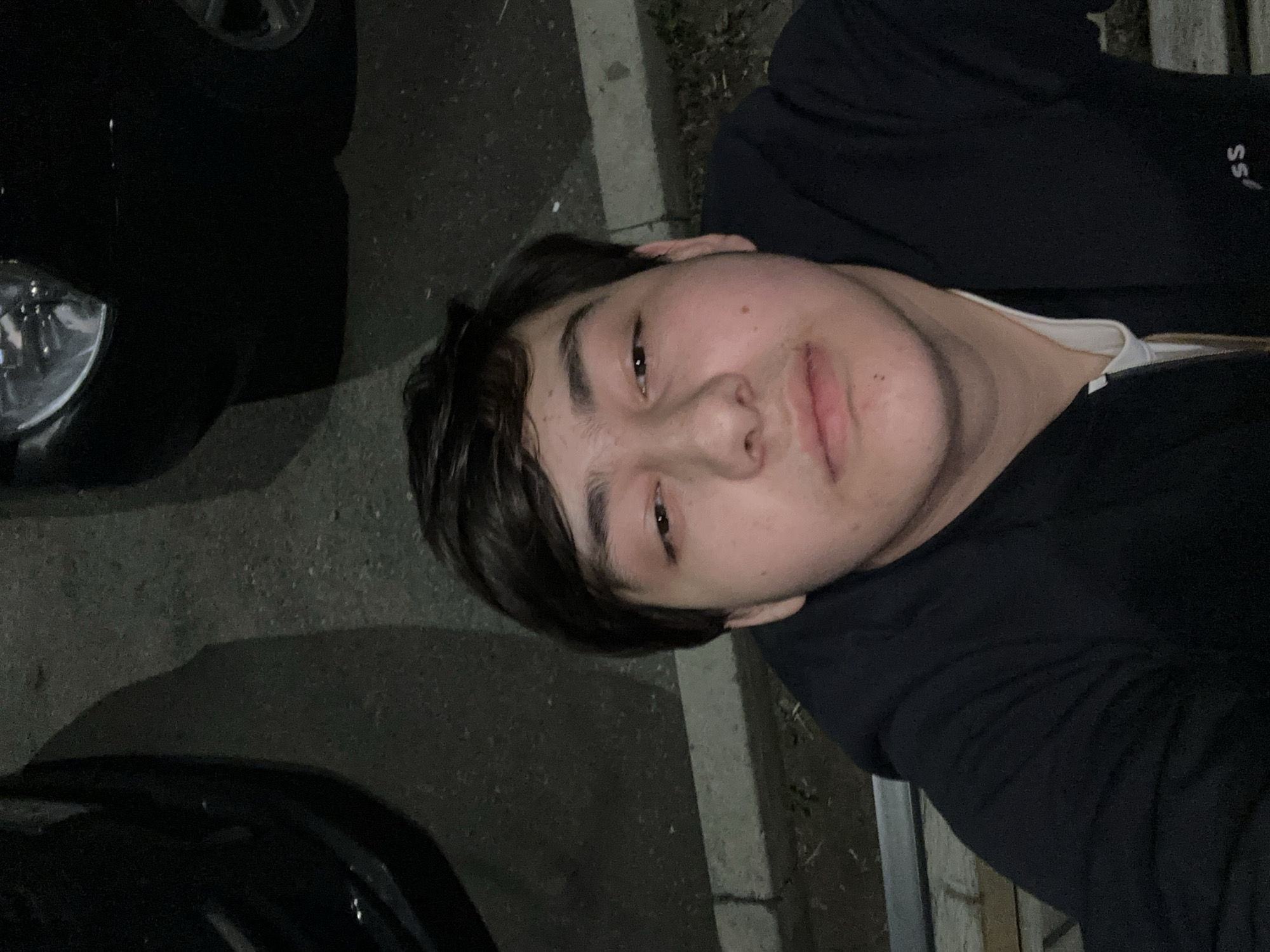 A young person is photographed outdoors at night in Unknown, Unknown. The person has short, dark hair and wears a black long-sleeved shirt over a white t-shirt. A black car, visible by its headlight and wheel, is positioned near the person. The ground appears to be paved, possibly a street or parking lot, and a concrete curb or barrier is visible next to the person.