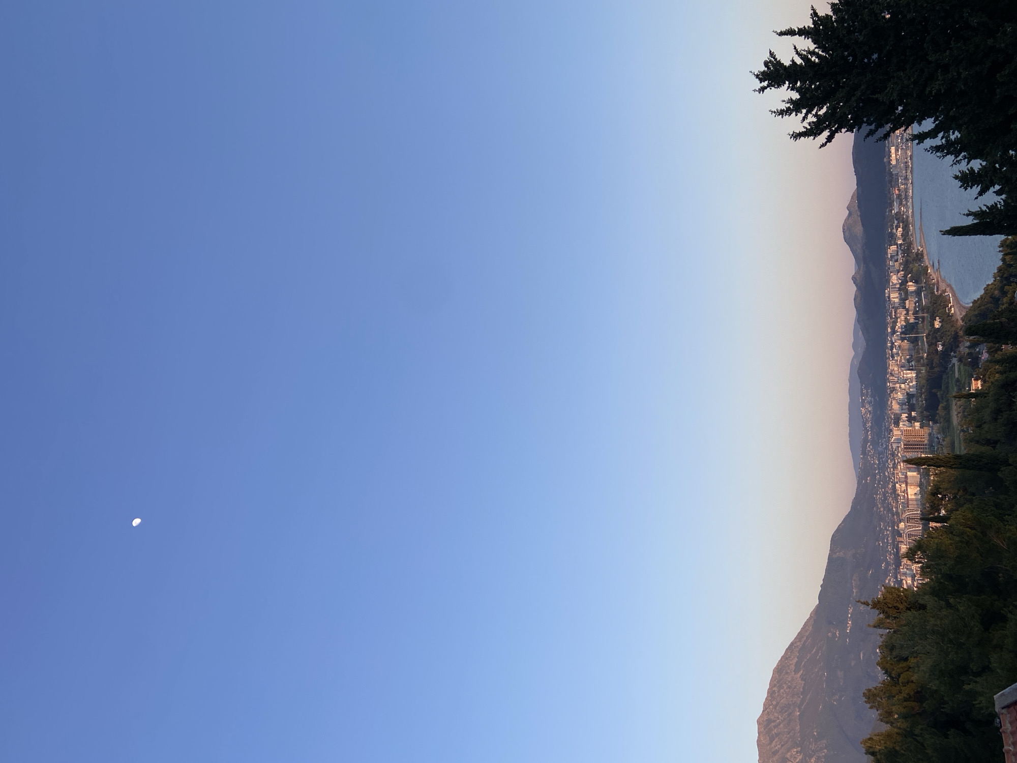 The image presents a landscape scene from the city of Šušanj, Montenegro. To the right, there's a mountain range with a city stretching along the coast, bordered by water. Trees frame the top right and bottom right edges of the image. To the left, a clear blue sky with a visible crescent moon is present. The horizon is illuminated by a subtle color gradient, suggesting either sunrise or sunset.