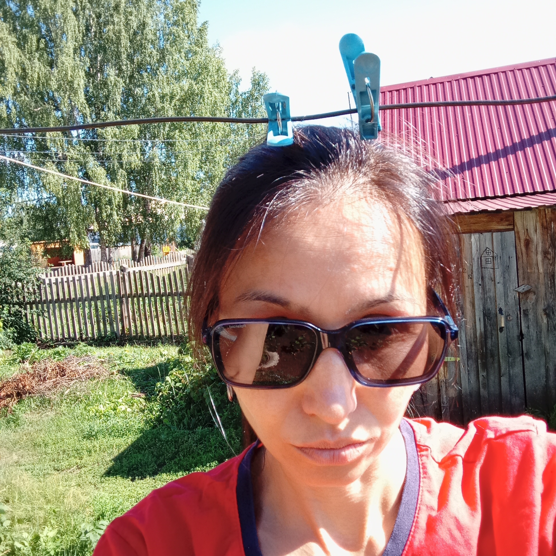 The image shows a woman wearing sunglasses and a red shirt, taking a selfie outdoors. Two blue clothespins are clipped to a string above her head. In the background, there is a wooden fence, green grass, trees, and a rustic wooden building with a red corrugated metal roof. The building appears to be a shed or outhouse, with a drawing of a house on its door. The scene is set in a rural location, likely a backyard or garden. The bright sunlight suggests a warm, clear day. The overall impression is one of a casual, outdoor setting in what is identified as Буренка, Russia.