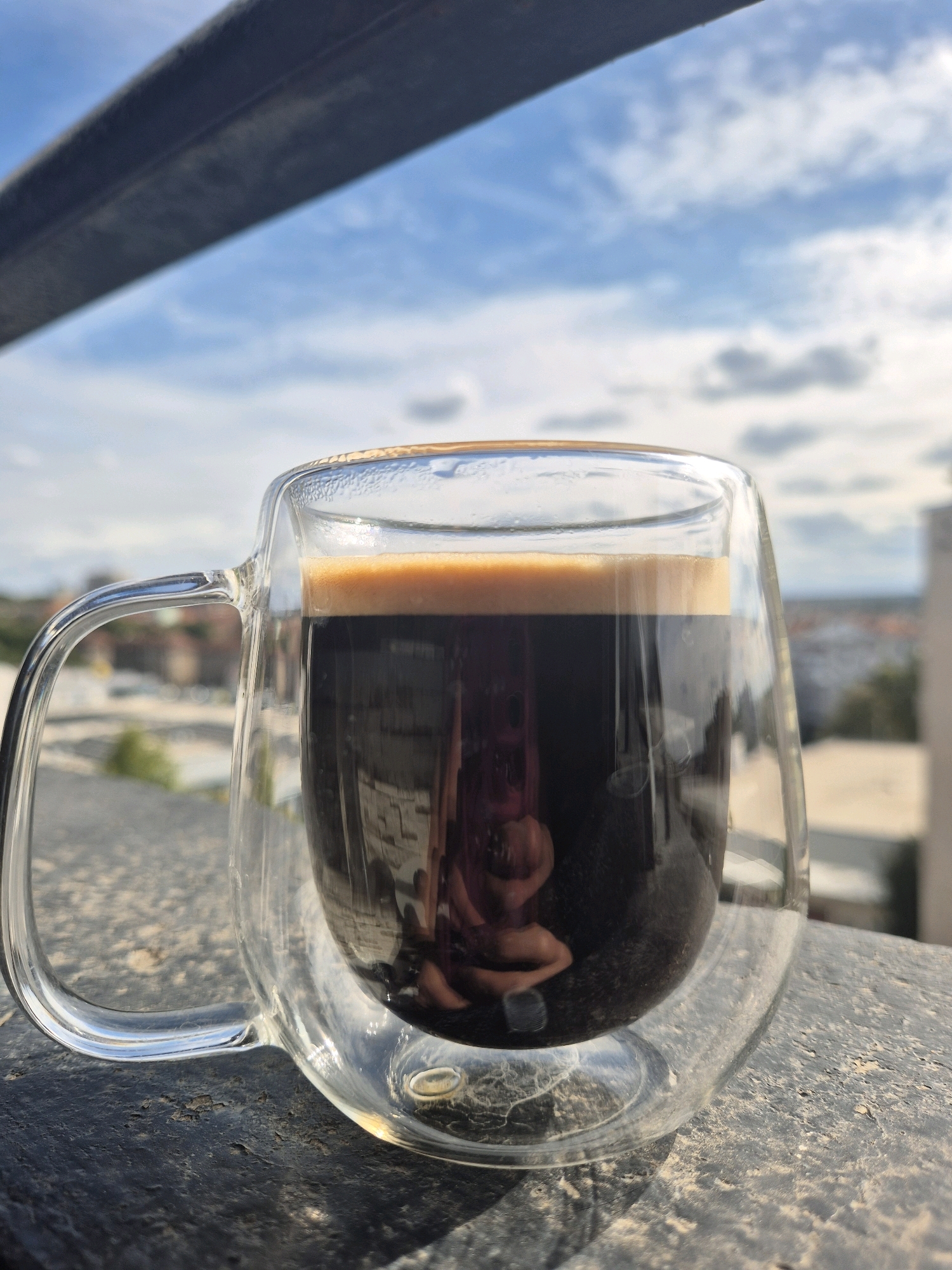 A close-up, eye-level shot shows a glass mug filled with dark espresso, topped with a layer of crema, sitting on a textured, dark surface. The mug has a clear handle and a double-walled glass construction. The background, slightly out of focus, reveals a cityscape with buildings and trees under a partly cloudy blue sky. A reflection of a person is visible in the coffee, hinting at the photographer's presence. A dark, horizontal beam or railing is positioned across the top of the frame. The lighting suggests it is daytime.