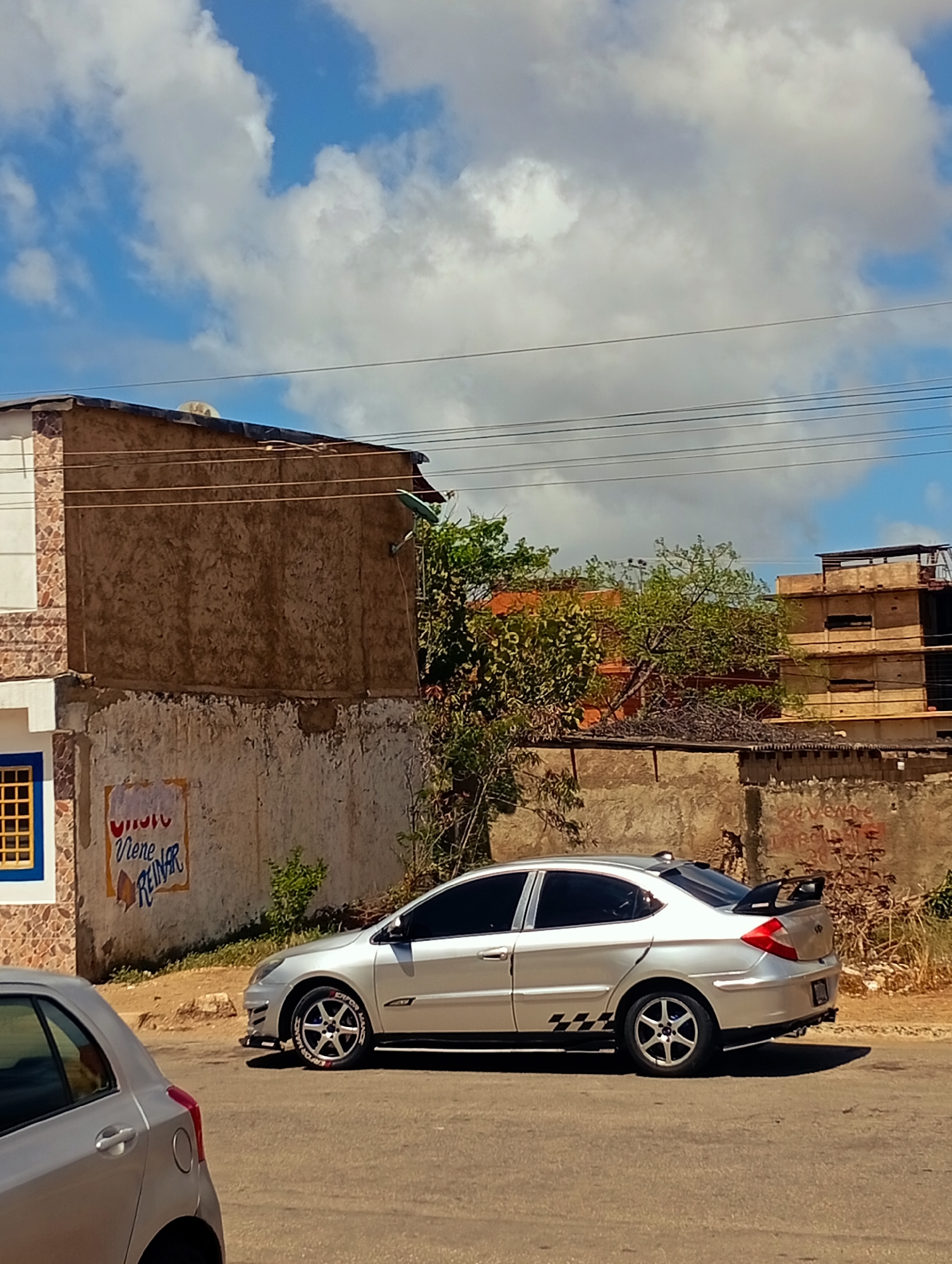 A silver sedan with a spoiler is parked on a street in Juan Griego, Venezuela. The car has black checkered racing stripes on its lower body and red lettering on its tires. A portion of another silver car is visible in the foreground to the left. Behind the parked car is a concrete wall with graffiti. Further back, there is a building with a textured brown facade, a white and patterned section on the left, and a blue-framed window. Beyond this, there are trees, more buildings, and power lines against a blue sky with white clouds. The ground is asphalt.