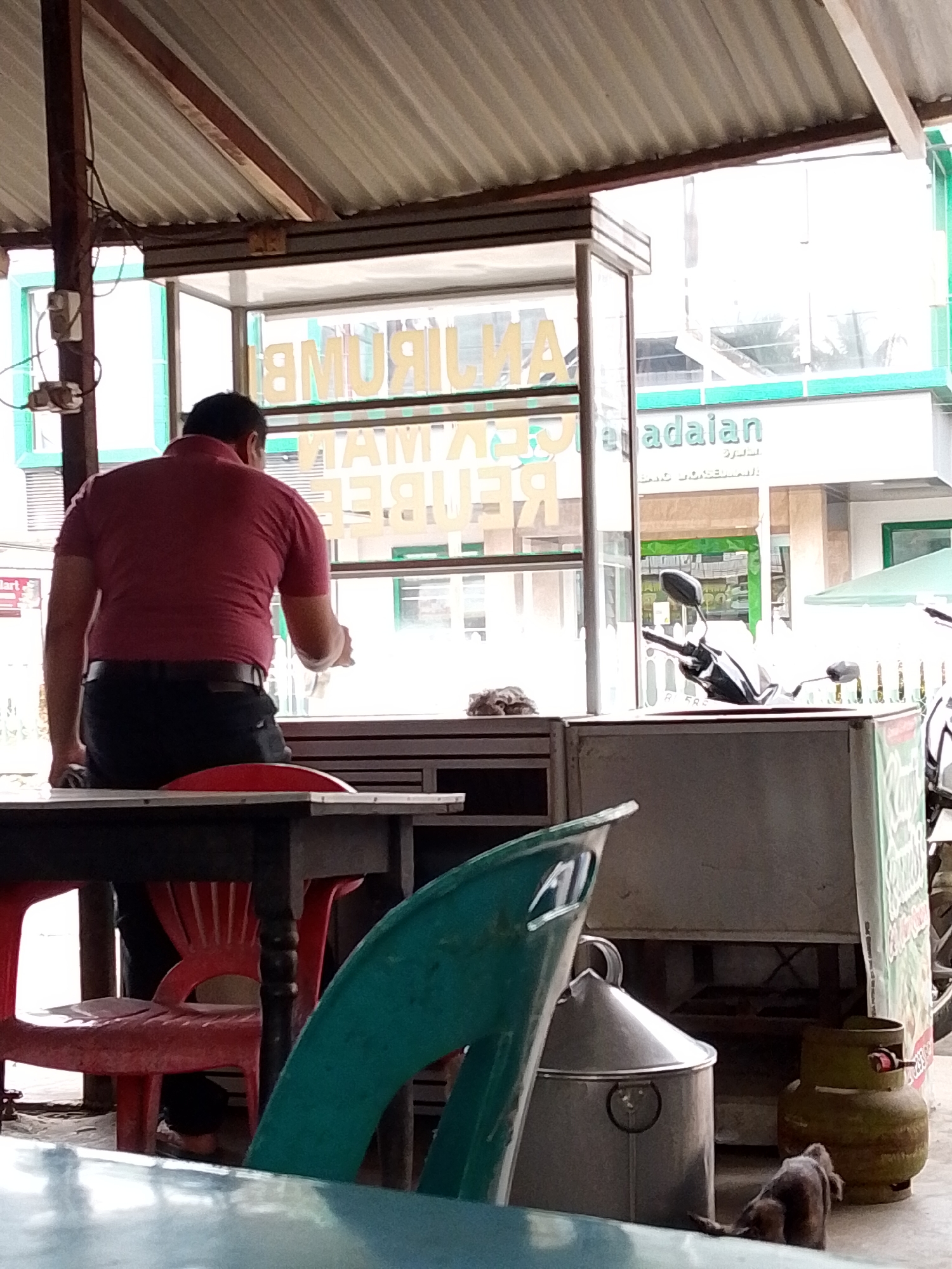 A man in a maroon polo shirt and dark pants is seated and turned away from the camera, facing a glass display case at an outdoor food stall. The stall appears to be located in Lhokseumawe, Indonesia, indicated by the text on the building in the background that reads "ADALAIAN". The man is holding a clear plastic cup in his right hand. In front of him, a red plastic chair is visible. The stall is covered by a corrugated metal roof supported by wooden beams and metal poles. Outside the stall, a motorcycle is parked. A gas cylinder and what appears to be a metal pot are on the ground near the stall. A small dog is visible in the lower right corner of the image, near the gas cylinder. The overall scene suggests a casual, everyday setting at a food vendor.