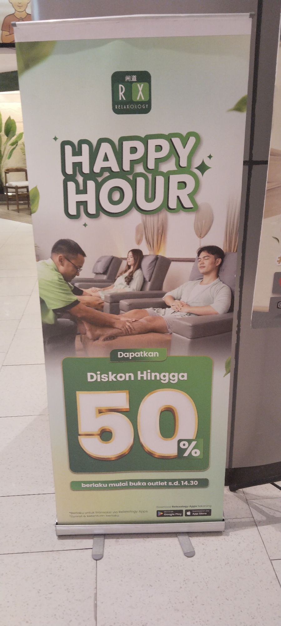 A vertical, outdoor banner advertising "Happy Hour" at Relaxology is displayed prominently. The banner features a green and white color scheme, with "HAPPY HOUR" emblazoned in large, bold, green letters across the top. Below the text, a photo depicts a foot massage being given to a person seated in a massage chair, with other individuals in similar chairs in the background. The bottom portion of the banner advertises a discount of "Diskon Hingga 50%" (Discount Up To 50%), with the percentage symbol rendered in a green square. Further down, smaller text indicates "berlaku mulai buka outlet s.d. 14.30" (valid from outlet opening until 14:30) and provides details on downloading the Relaxology app from Google Play and the App Store. The banner is supported by two silver metal legs and appears to be set up in a public area, possibly a mall or shopping center, with a tiled floor visible at the bottom. The context of Semarang, Indonesia, is not directly evident from the visual information presented in the image itself, but this location is provided as a prerequisite for the description.