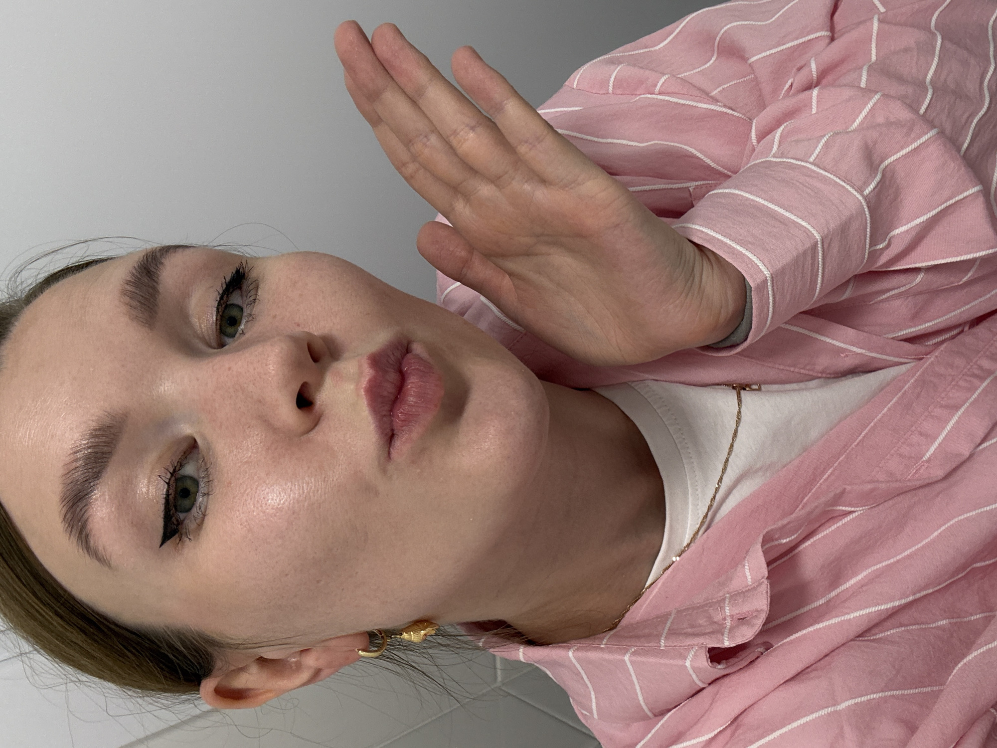 A young woman with fair skin, light brown hair, and green eyes is taking a selfie in what appears to be a bathroom. She is wearing a pink and white striped button-down shirt over a white t-shirt, a gold necklace, and a gold hoop earring with a seashell charm. Her makeup includes winged eyeliner and lip gloss. She is puckering her lips and extending her left hand towards the camera as if blowing a kiss. The background shows a white tiled wall and a gray wall. There is no identifiable information in the image that can link it to Izhevsk, Russia.