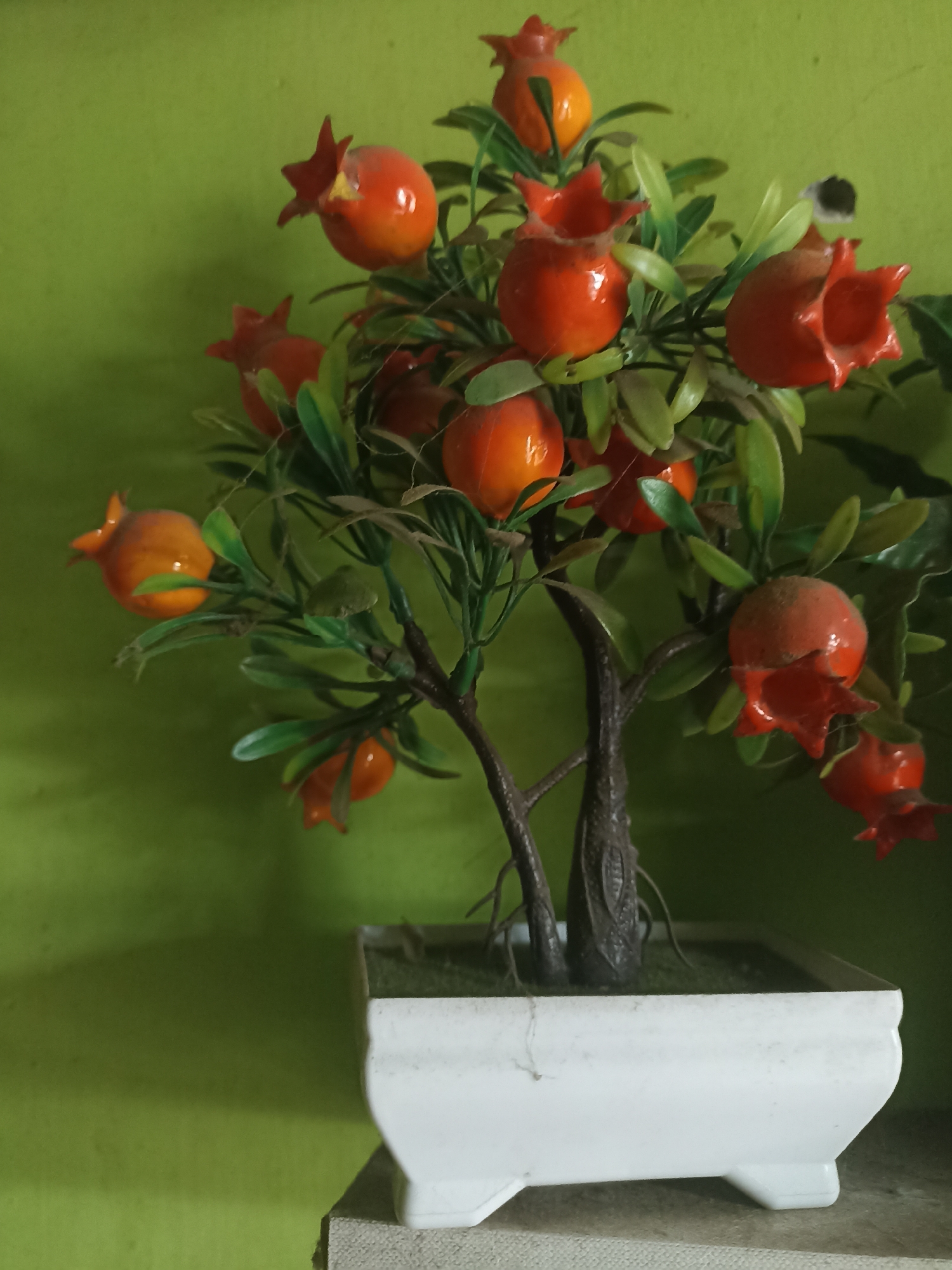 The image shows a close-up of an artificial pomegranate tree in a white pot. The tree has green leaves and several orange-red pomegranates. The pot is square with rounded edges and sits on a light-colored surface. The background is a solid, textured lime green wall. There are no people visible in the image, and no actions or interactions are taking place. The setting appears to be indoors. There is no information to suggest this image is located in Pandanarum, Indonesia.