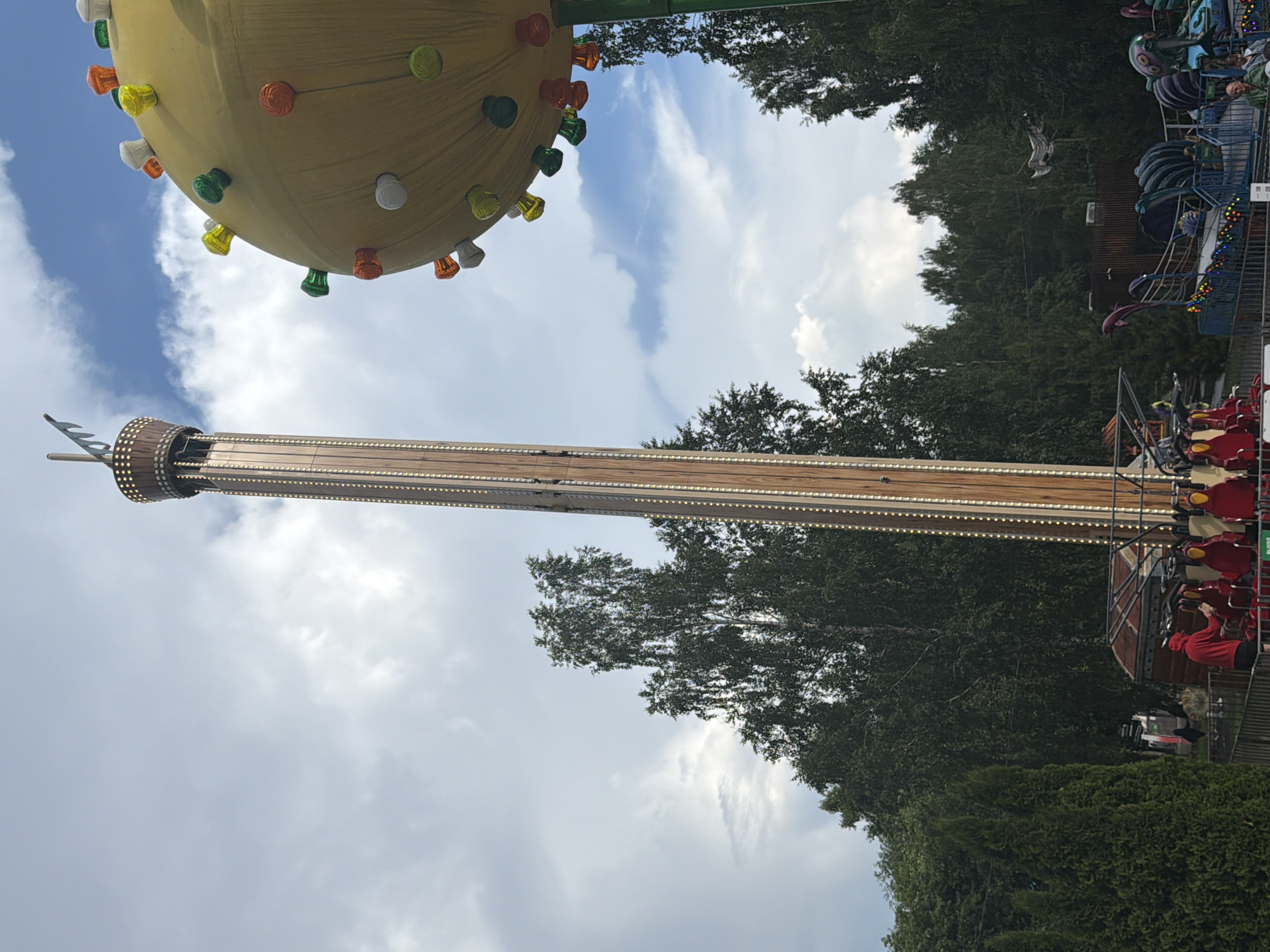 An outdoor shot captures an amusement park ride, a tall, slender tower structure with a wooden facade adorned with lights, standing against a cloudy sky. To the right, a large yellow spherical object, also decorated with colorful lights, is partially visible. In the foreground, several red seats with yellow restraints are arranged, with a person in a red hoodie visible near them, suggesting the ride is either about to begin or has just concluded. The background features a dense line of dark green trees, indicating the park is situated amidst natural surroundings. Further back, other amusement park rides can be seen, including one with a dolphin-like sculpture and a ride with octopus-like arms. The overall scene depicts a lively day at an amusement park.
