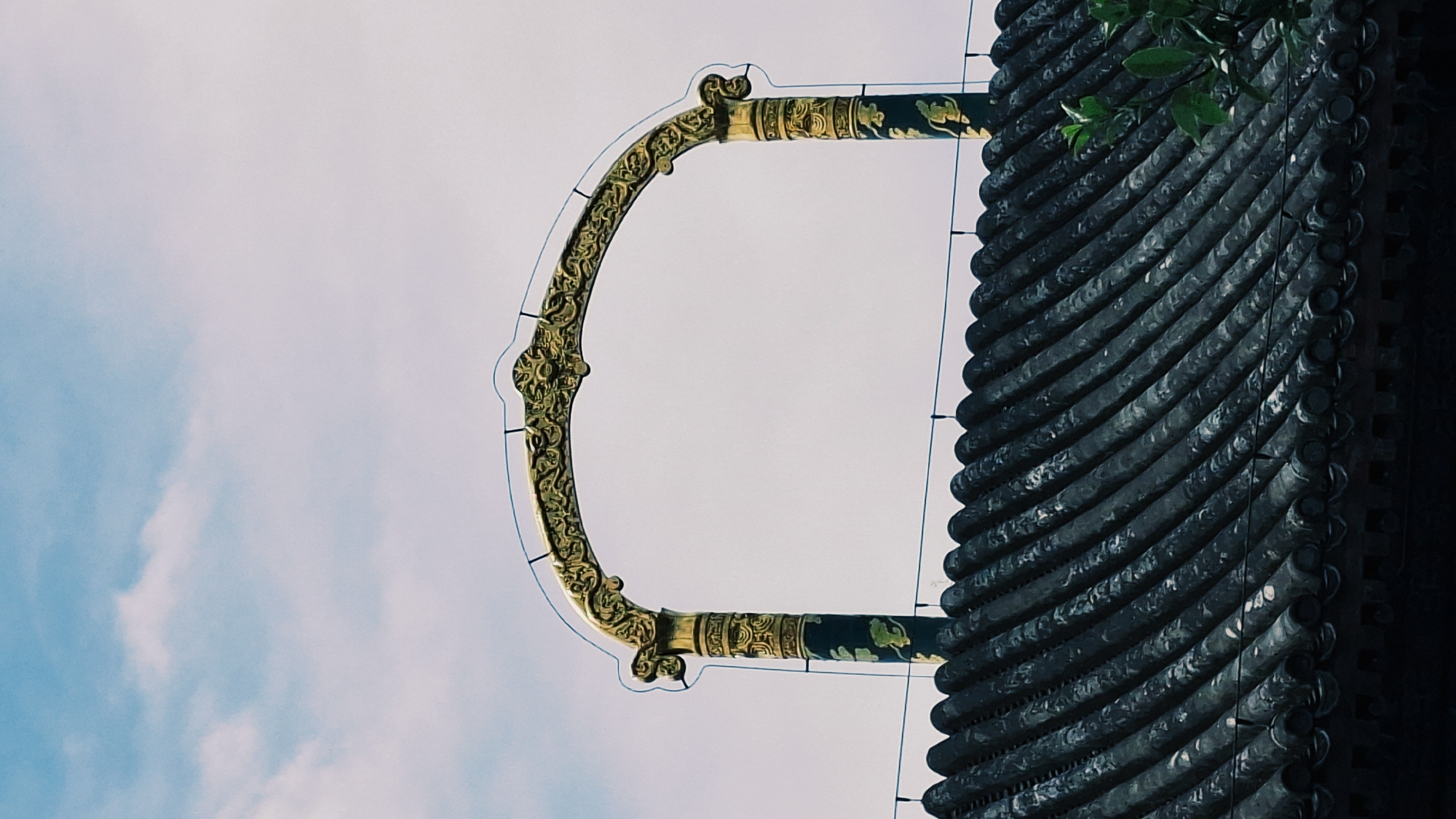 From a low angle, the image shows a segment of an ornate Chinese building. The building's dark-colored roof tiles are the most prominent feature, cascading in horizontal rows. A decorative, semi-circular, gold-accented bracket extends from the roof, reaching toward the sky, with a blue and white sky backdrop. The location is likely in Haidian District, China, given the architectural style, although there is no explicit confirmation.
