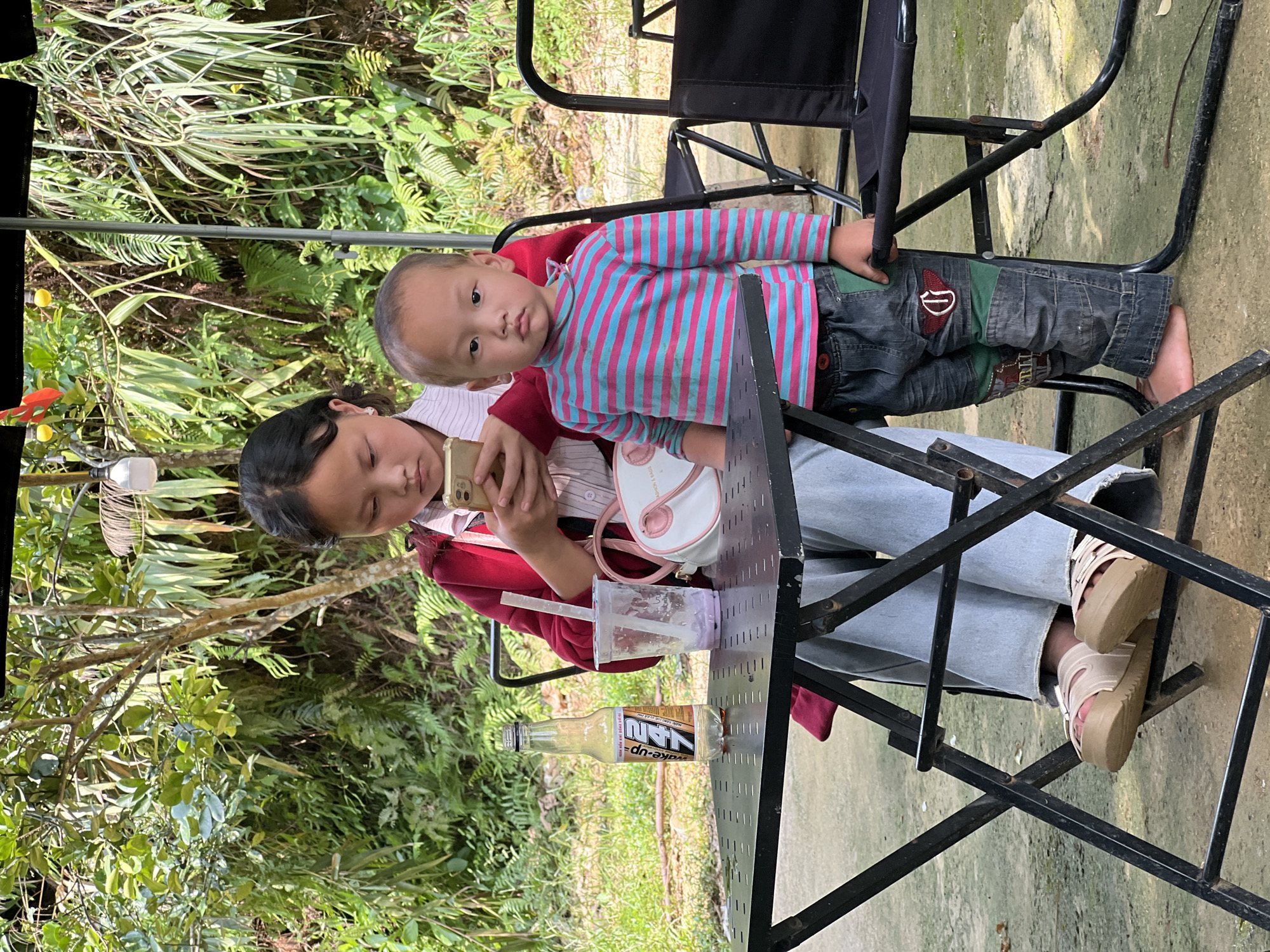 In Lai Dong, Vietnam, two children are seated at a metal folding table outdoors. A young girl on the left, wearing a red jacket and light blue jeans, is looking at her phone. Beside her is a plastic cup with a straw and a bottle of 247 energy drink. A young boy on the right, dressed in a striped long-sleeve shirt and patched jeans, stands and looks directly at the camera. The background features lush green foliage, indicating a natural or park-like setting.