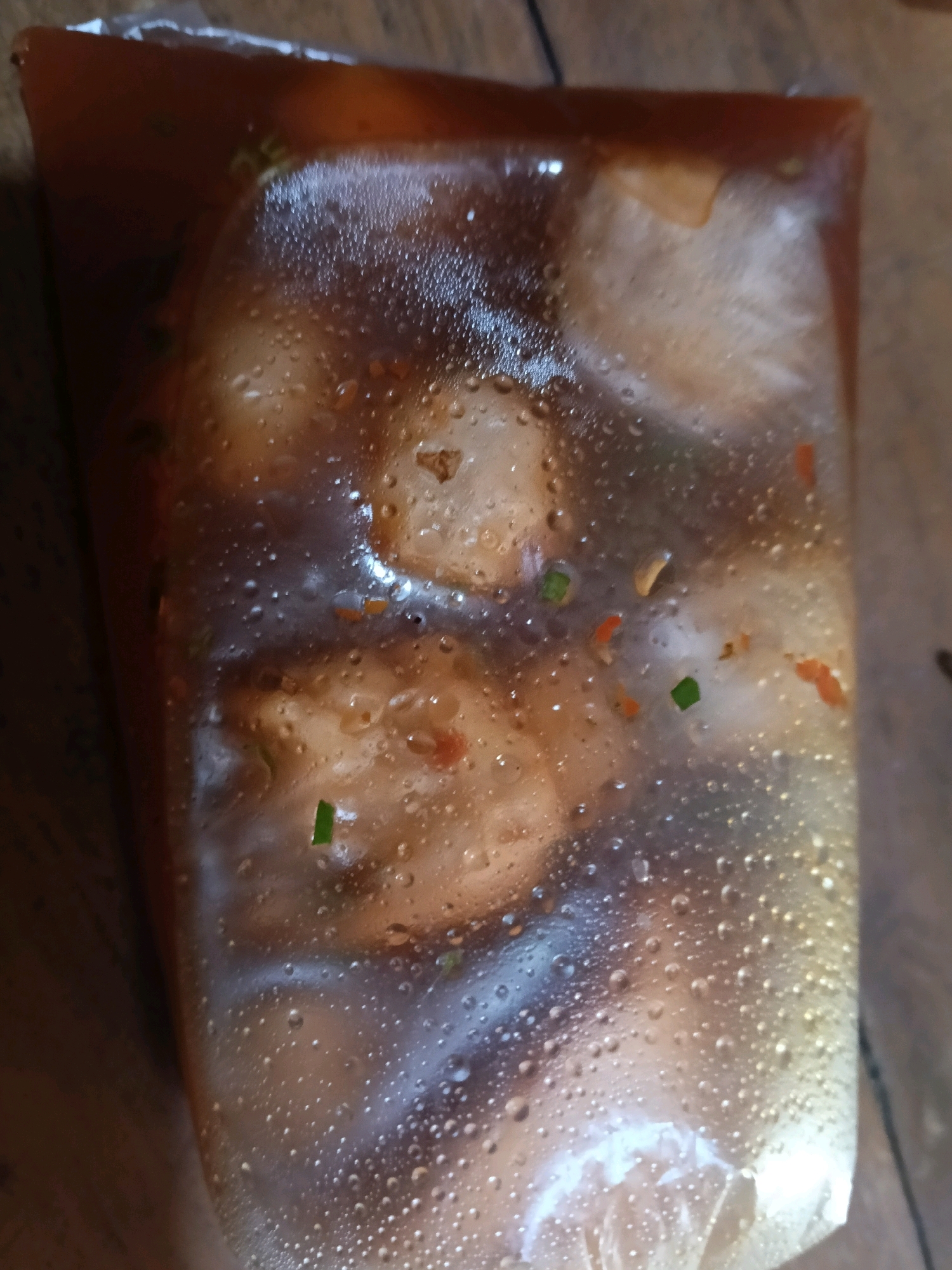 The image shows a close-up, vertical shot of a clear plastic bag filled with food. The food appears to be meatballs or dumplings in a dark, savory sauce. Small droplets of condensation are visible on the inside of the plastic bag, indicating the food might be warm or was recently refrigerated. Some red chili flakes and green scallions are also scattered throughout the bag. The plastic bag is sealed at the top. The background is a blurry, brown surface that appears to be a wooden table or countertop. There are no people or explicit actions depicted. The image does not contain any information or context about the city of Nglungger, Indonesia.
