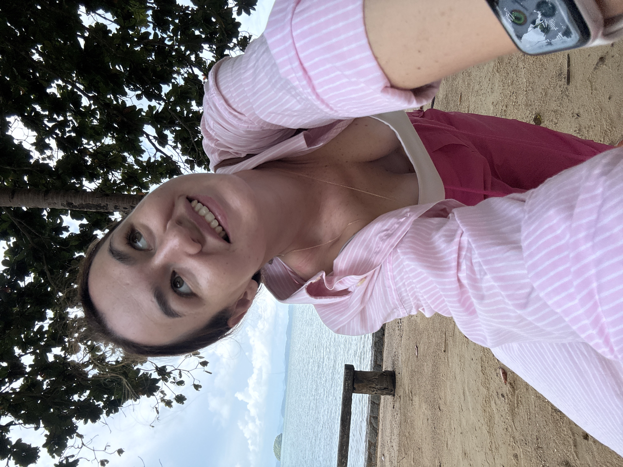 A woman is taking a selfie while crouching on a sandy beach in Pa Khlok, Thailand. She is wearing a pink and white striped button-down shirt over a white top and pink pants. The ocean and some distant islands are visible in the background, along with a wooden bench and a tree with dark green leaves overhead. The sky is overcast with clouds.