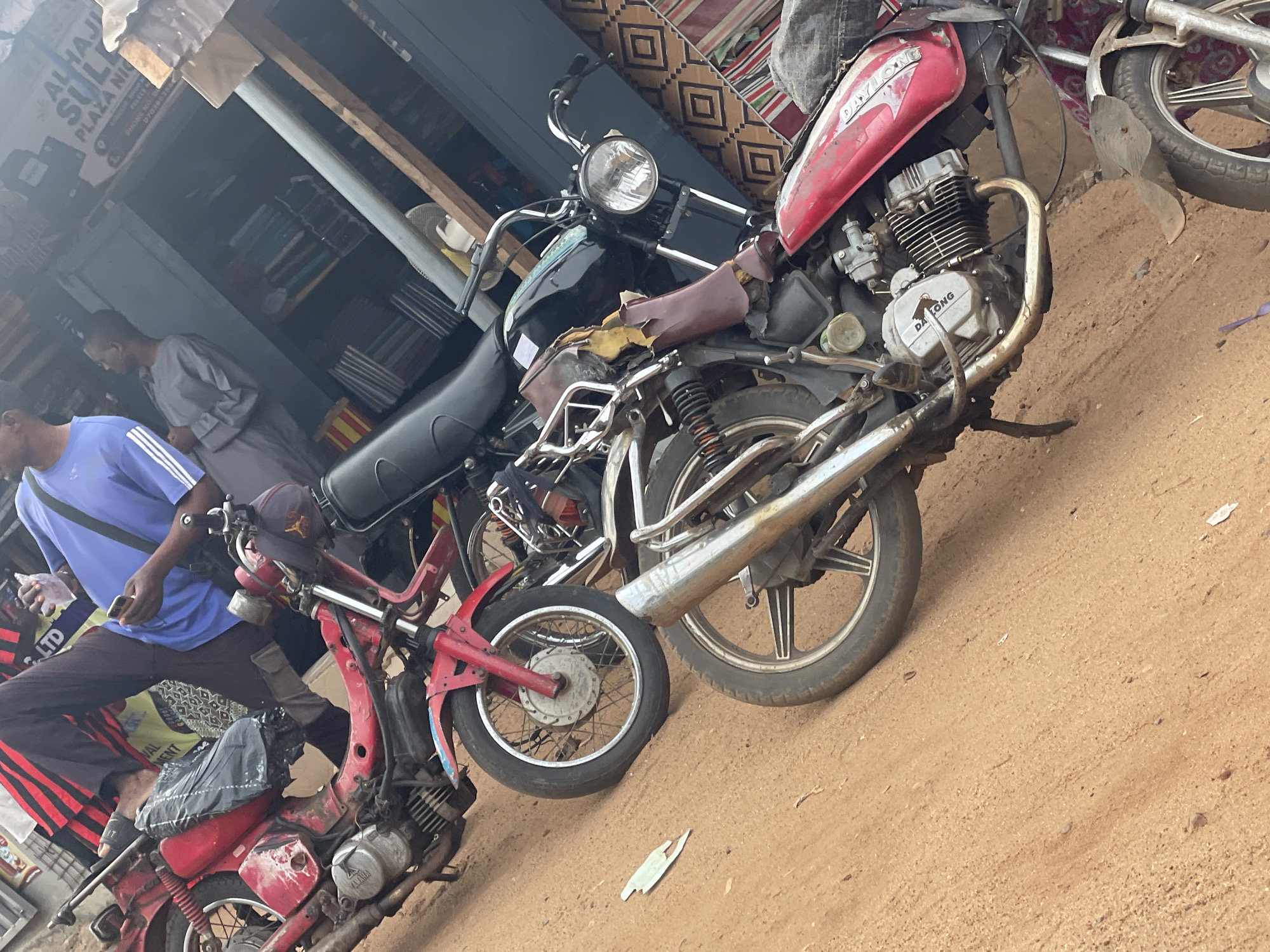 In Lokoja, Nigeria, two motorcycles are parked on a dirt road in front of a shop. A red motorcycle with visible wear and tear is in the foreground, its engine exposed. Behind it, a black motorcycle is partially visible. Two men are present; one in a blue shirt holds a phone, and another in a grey robe stands nearby. The shop behind them displays goods and signage that is partially obscured. The scene appears to be a typical street view in a Nigerian city, characterized by the vehicles, the open-air shop, and the unpaved ground.