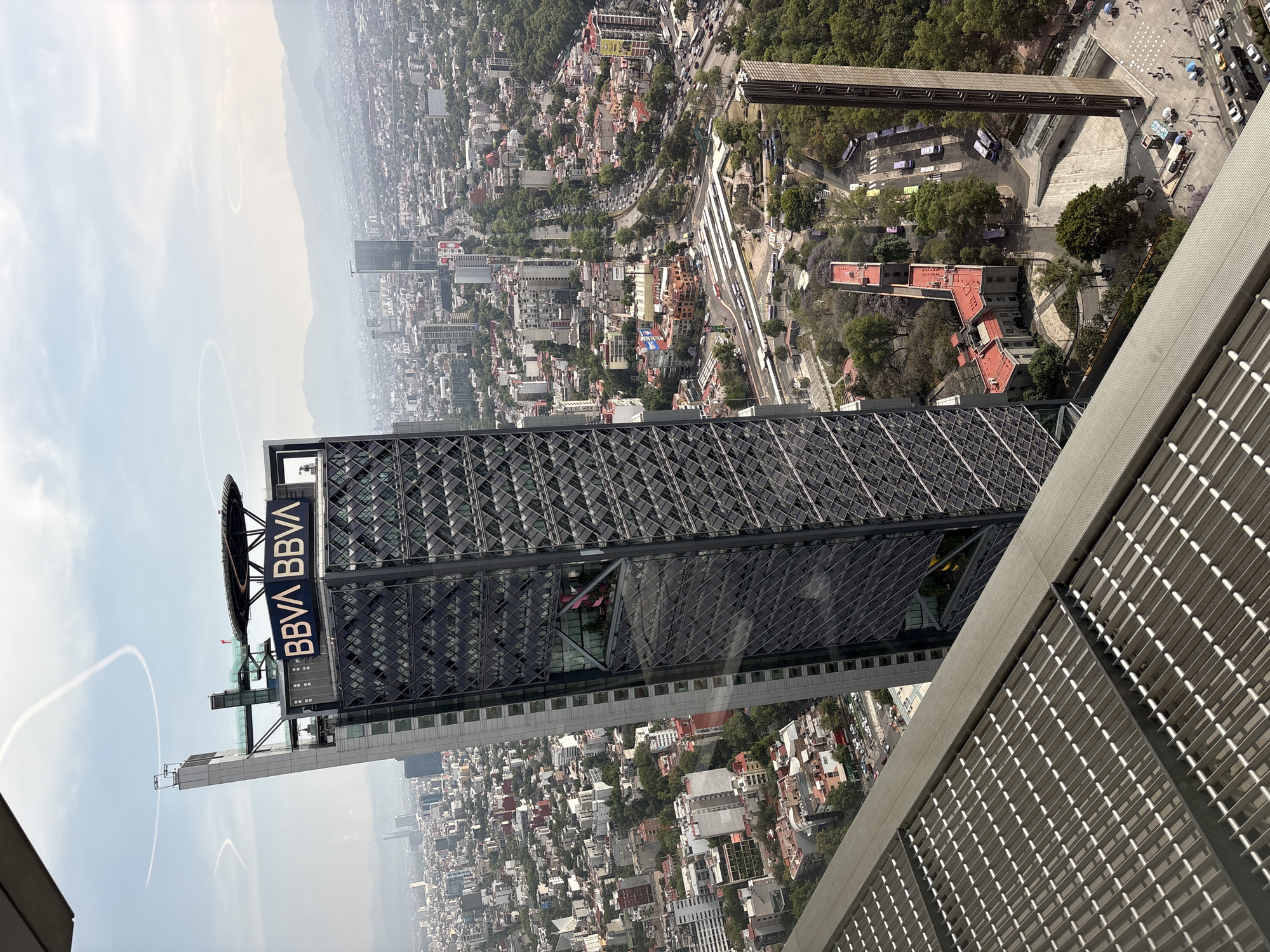 The image shows a high-angle view of the BBVA tower in Mexico City, Mexico. The tower is a prominent feature of the cityscape and features a dark gray geometric facade with the "BBVA" logo prominently displayed on its upper portion. The building appears to be viewed from a nearby skyscraper, showing a section of the building's exterior, which includes a grid-like metal structure. Below the buildings are several other buildings, highways, and trees. The sky is visible to the left of the image, showing white contrails in the clear blue sky.