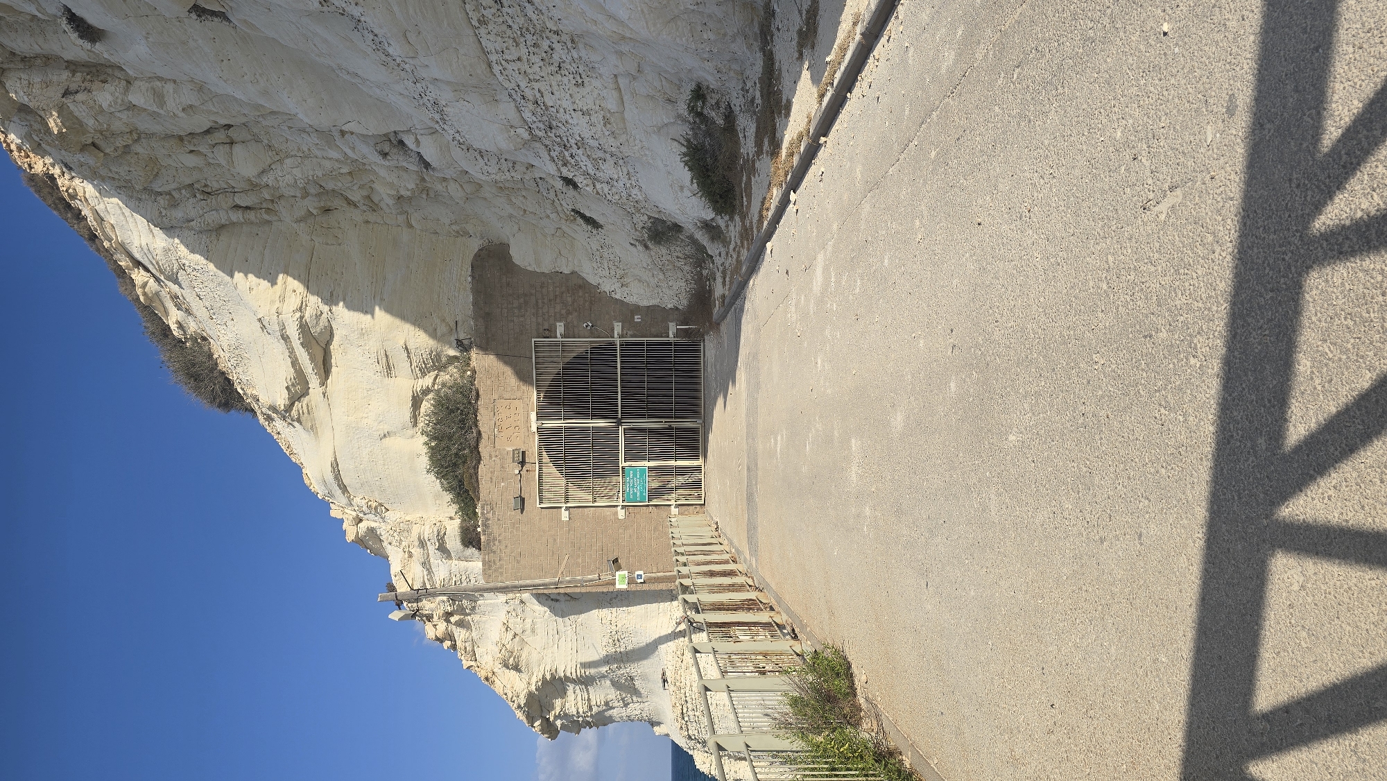 The image shows a paved pathway leading to a large, barred entrance set into a white, rocky cliff face. The entrance is made of metal bars and has a green sign with text on it. There are lights mounted on the brick wall surrounding the entrance. A metal fence runs along the left side of the pathway, separating it from a grassy area and the sea. The cliff face is sheer and layered, with some vegetation growing on top. The sky is clear and blue, and the sun casts a shadow from the fence onto the pathway. The location is identified as Rosh HaNikra grottoes in the Mate Asher Regional Council, Israel. There are no people visible in the image.
