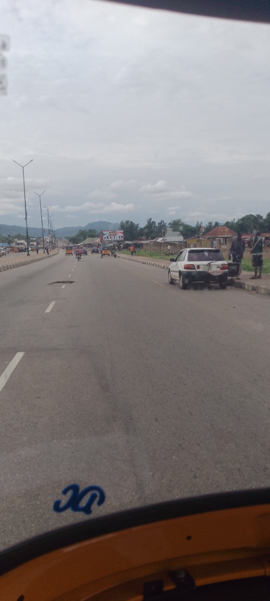 The image shows a wide, paved road in Jalingo, Nigeria, with a cloudy sky overhead. The road has multiple lanes and is marked with white lines. There are streetlights visible along the side of the road. In the distance, there are buildings, trees, and hills. Several vehicles are present on the road, including cars, motorcycles, and what appears to be an auto-rickshaw. Some people are visible standing on the side of the road and near the vehicles. One car, a white sedan, is parked on the side of the road with two men standing nearby. The road surface shows signs of wear, including some potholes. The foreground of the image shows a portion of a yellow vehicle, possibly the one from which the photo was taken, with blue lettering on its surface. The overall scene depicts a typical urban street environment in Jalingo.