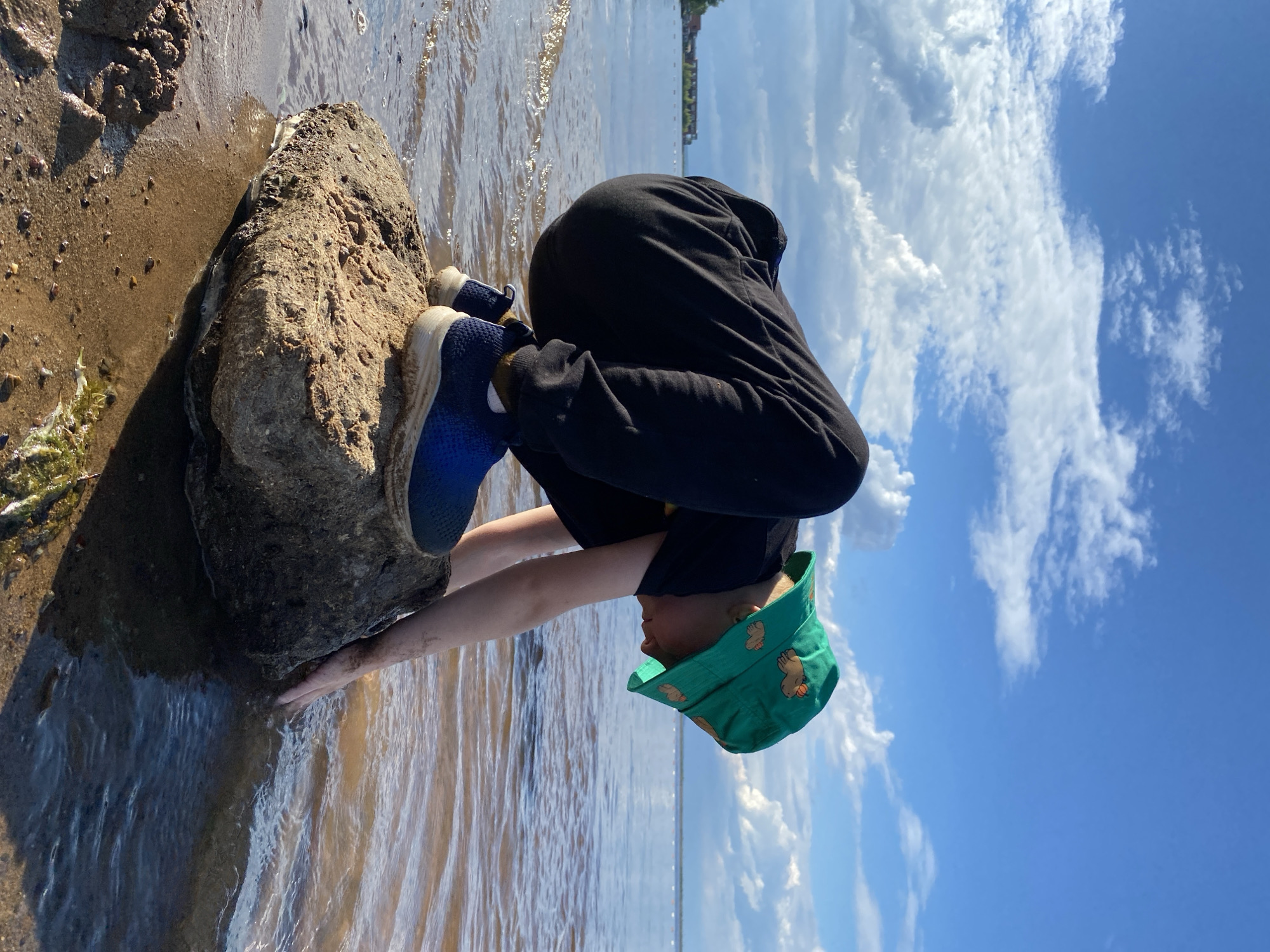 A child wearing a green bucket hat with cartoonish animal designs and black pants crouches on a large, wet rock by a body of water. The child's right hand is in the water, and their left hand is resting on the rock. The child's face is partially obscured, but they appear to be looking down at the water. Blue sneakers are visible on the child's feet. The environment includes a sandy shoreline with some vegetation, a calm body of water with ripples, and a cloudy blue sky. In the distance, across the water, there is a faint industrial-looking structure with a line of trees. The scene is likely located at the Kama River embankment in Chaikovsky, Russia, given the industrial structure and the wide river.