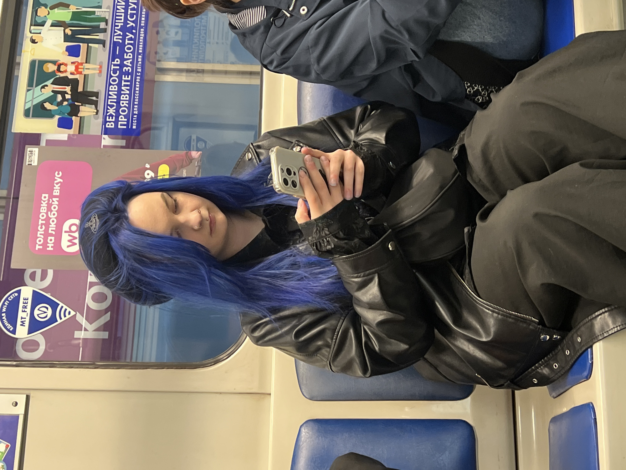 A woman with dyed blue hair is sitting on a blue seat in the Saint Petersburg, Russia metro while looking at her cell phone. She is wearing a black leather jacket and black pants. Next to her, another person is also sitting. Advertisements are visible on the train window.