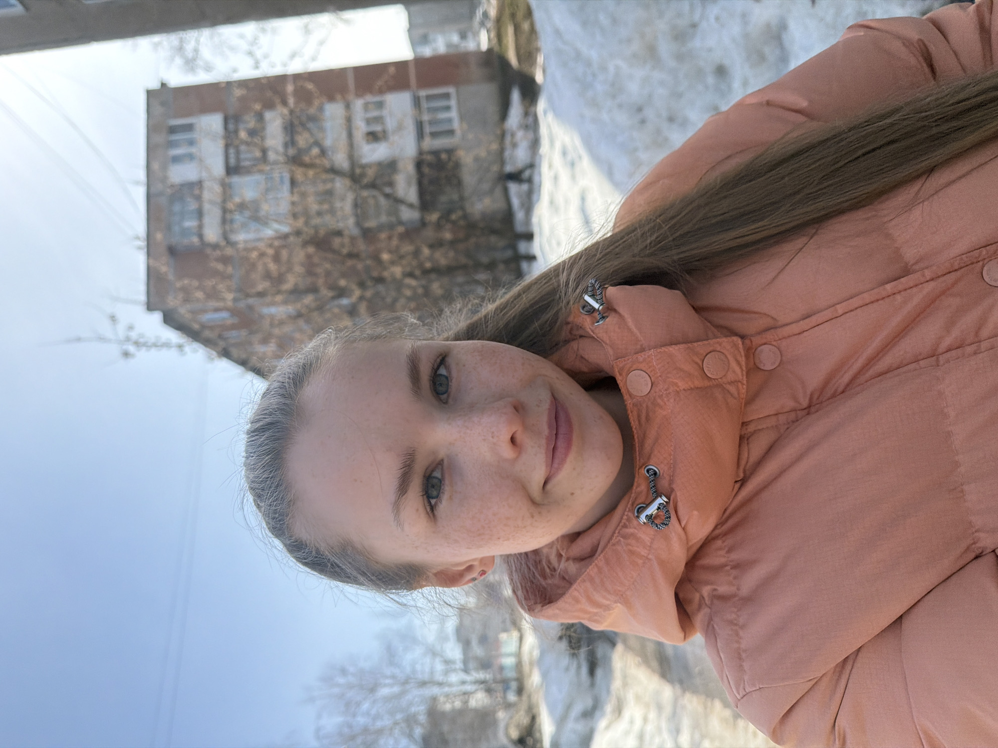 A young woman is positioned outdoors, likely taking a selfie. She's wearing an orange puffer jacket. Her hair is pulled back. Freckles are visible on her face. The background includes a multi-story building with windows. Snow is visible on the ground, and bare trees are present. The location is in Berezniki, Russia.