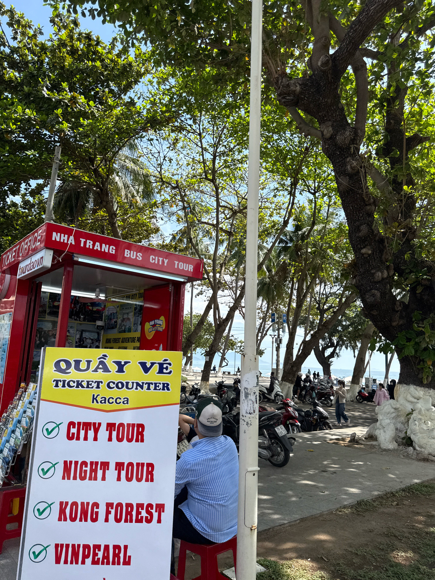 In Nha Trang, Vietnam, a bright red ticket booth with "NHA TRANG BUS CITY TOUR" and "TICKET OFFICE" signage is positioned next to a sidewalk shaded by large trees. In front of the booth, a yellow and white sign lists available tours: "CITY TOUR," "NIGHT TOUR," "KONG FOREST," and "VINPEARL," all with green checkmarks. A person wearing a blue and white striped shirt and a baseball cap is seated on a red stool to the right of the sign, facing away from the camera. Behind the booth, a sidewalk lines the beach, with several motorcycles parked along it. In the background, people are visible near the ocean and trees, with palm trees interspersed amongst the larger deciduous trees. The sky is clear and blue.