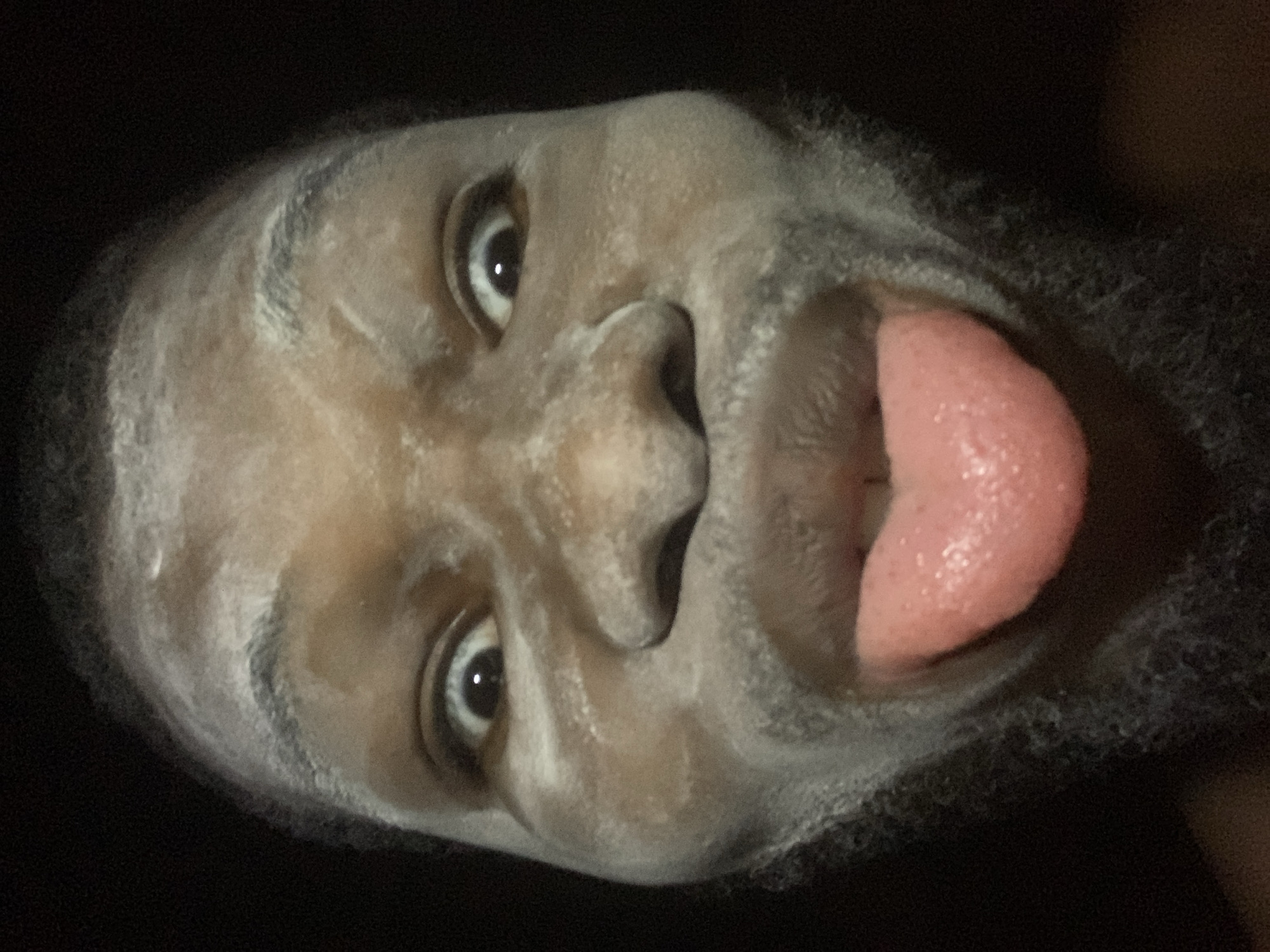 A close-up, slightly low-angle, and poorly illuminated photograph shows the face of a Black male with a beard. He has white powder or residue on his face and hair, and his tongue is sticking out. The background is dark and indistinct. There is no contextual information to indicate the location in Onitsha, Nigeria.