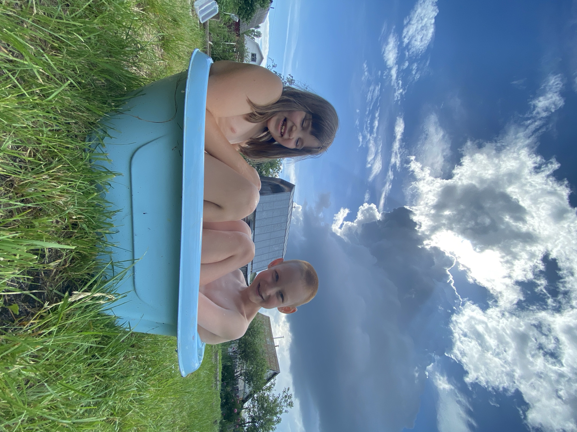 Two children, a girl and a boy, are sitting in a blue plastic tub filled with water, likely a small inflatable pool. The girl, with long brown hair, is positioned behind the boy, her torso visible as she leans back and smiles widely. The boy, with short red hair, is in front and facing the camera, also smiling. Both appear to be undressed from the waist up, suggesting they are bathing or playing in the water.
The tub is situated outdoors on a grassy area, with tall green grass surrounding it. In the background, hints of a rural or suburban setting are visible, including houses with roofs and some trees. The sky above is a prominent feature, filled with dramatic clouds, ranging from bright white and fluffy to darker, more ominous storm clouds. Sunlight breaks through in some areas, suggesting a partly cloudy day, possibly with a storm approaching or passing.
The overall scene conveys a sense of fun and childhood enjoyment, with the children appearing happy and engaged in their activity. The lighting suggests it is daytime, likely afternoon. The provided location context is Chaikovsky, Russia. No visible text is present in the image.