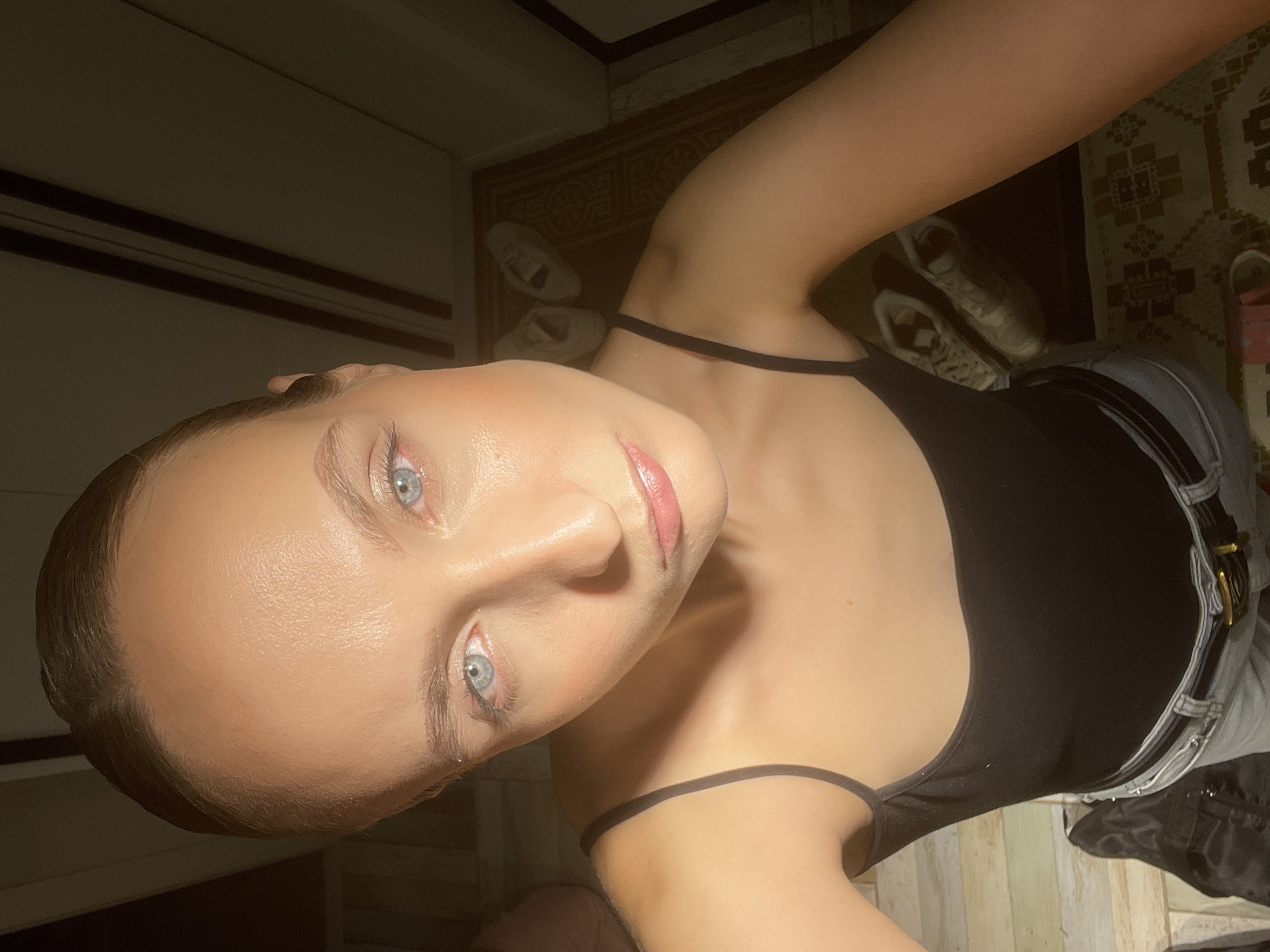 A young woman with light skin and blue eyes is shown in a close-up, overhead shot. She has her hair pulled back and is wearing a black spaghetti strap top and light-wash jeans with a belt. The lighting is dramatic, with a strong light source from the top right casting shadows. In the background, a rug with a geometric pattern is visible, along with a pair of white sneakers and what appears to be wooden flooring or paneling. The overall setting suggests an indoor environment, possibly a home. The location is identified as Chaikovsky, Russia.