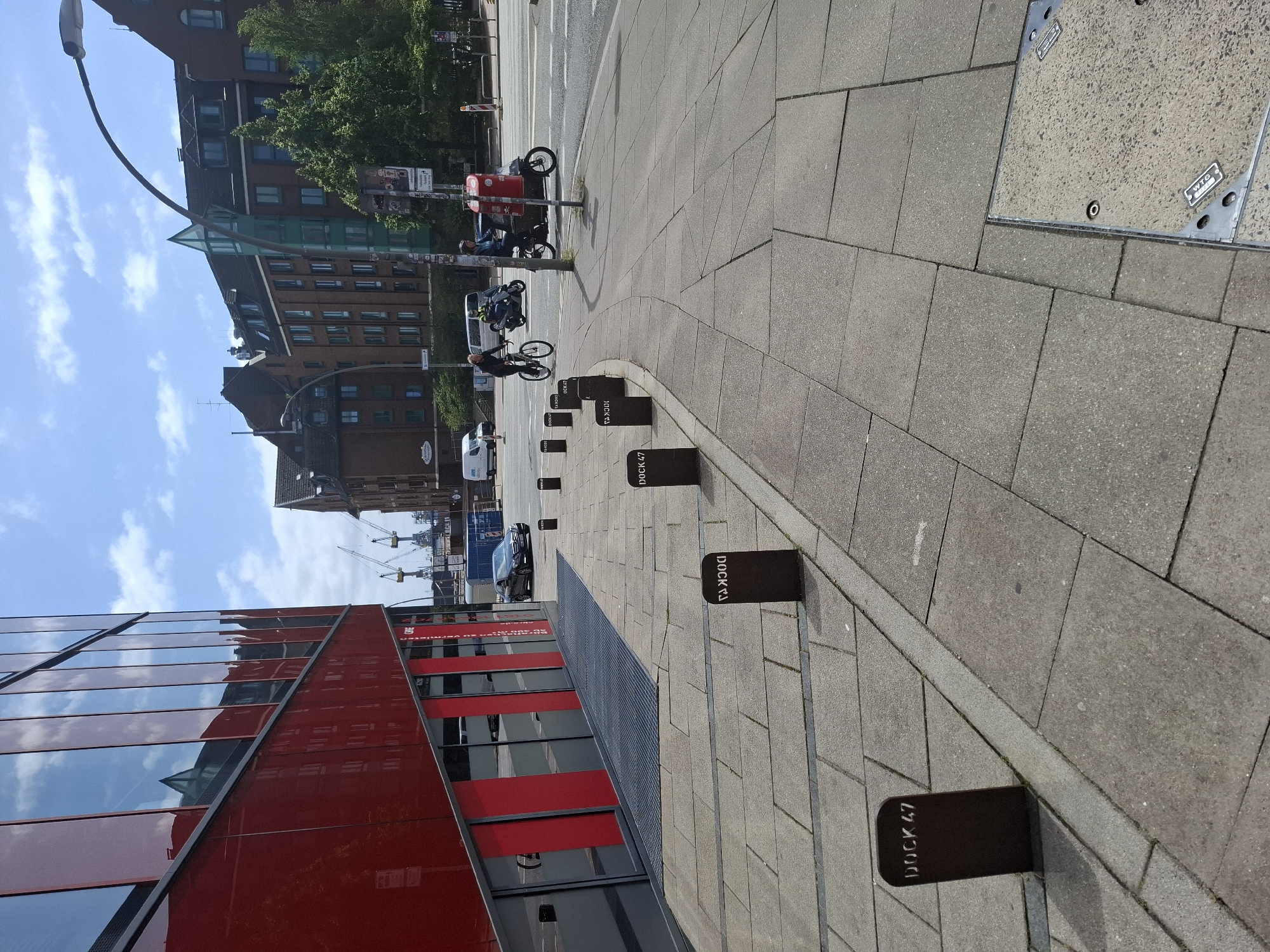 The image shows a street scene in Hamburg, Germany, specifically at Dock 47 in the city's harbor district. In the foreground, a paved sidewalk is lined with dark, rectangular bollards, each stenciled with "DOCK 47". The sidewalk is adjacent to a modern building with a striking red facade and large, reflective windows. Beyond the sidewalk, a street is visible with vehicles including a black Mercedes sedan, a bicycle, and a scooter. Pedestrians are also present on the sidewalk. In the background, traditional brick buildings and industrial cranes suggest the area's maritime history. The sky is blue with scattered clouds.