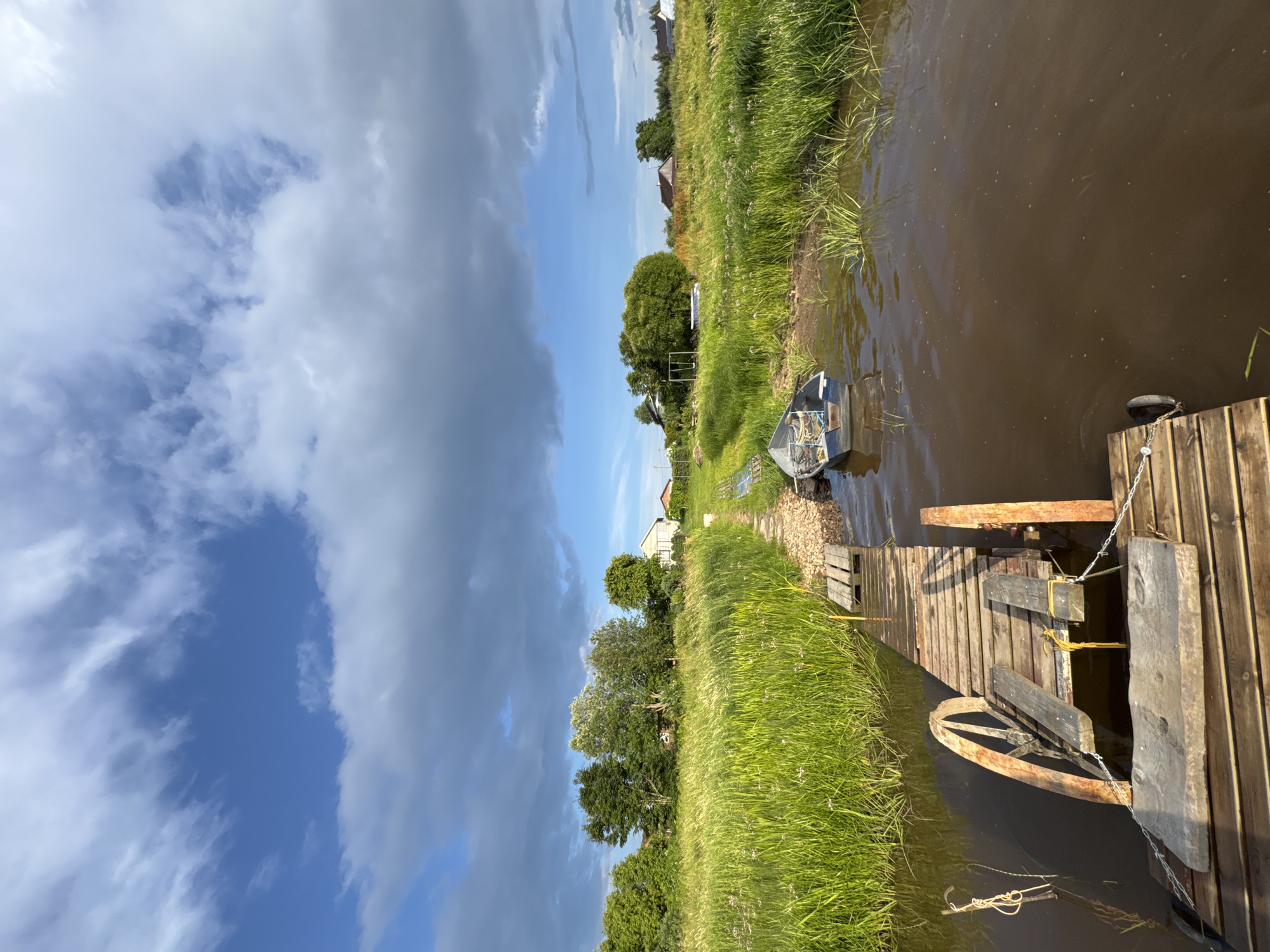 The image depicts a serene rural landscape in Sitno, Russia. In the foreground, a weathered wooden pier extends into murky brown water. A small, weathered blue boat is moored to the pier, facing away from the viewer. The pier is made of planks and has some visible signs of age and wear. Beyond the pier, a path of gravel and grass leads towards a lush green field filled with tall grass and scattered wildflowers. In the background, several houses with traditional Russian architecture are nestled amongst trees. The sky above is a mix of vibrant blue and dramatic gray clouds, hinting at a changing weather pattern. The overall impression is one of peaceful, rustic, and natural beauty.