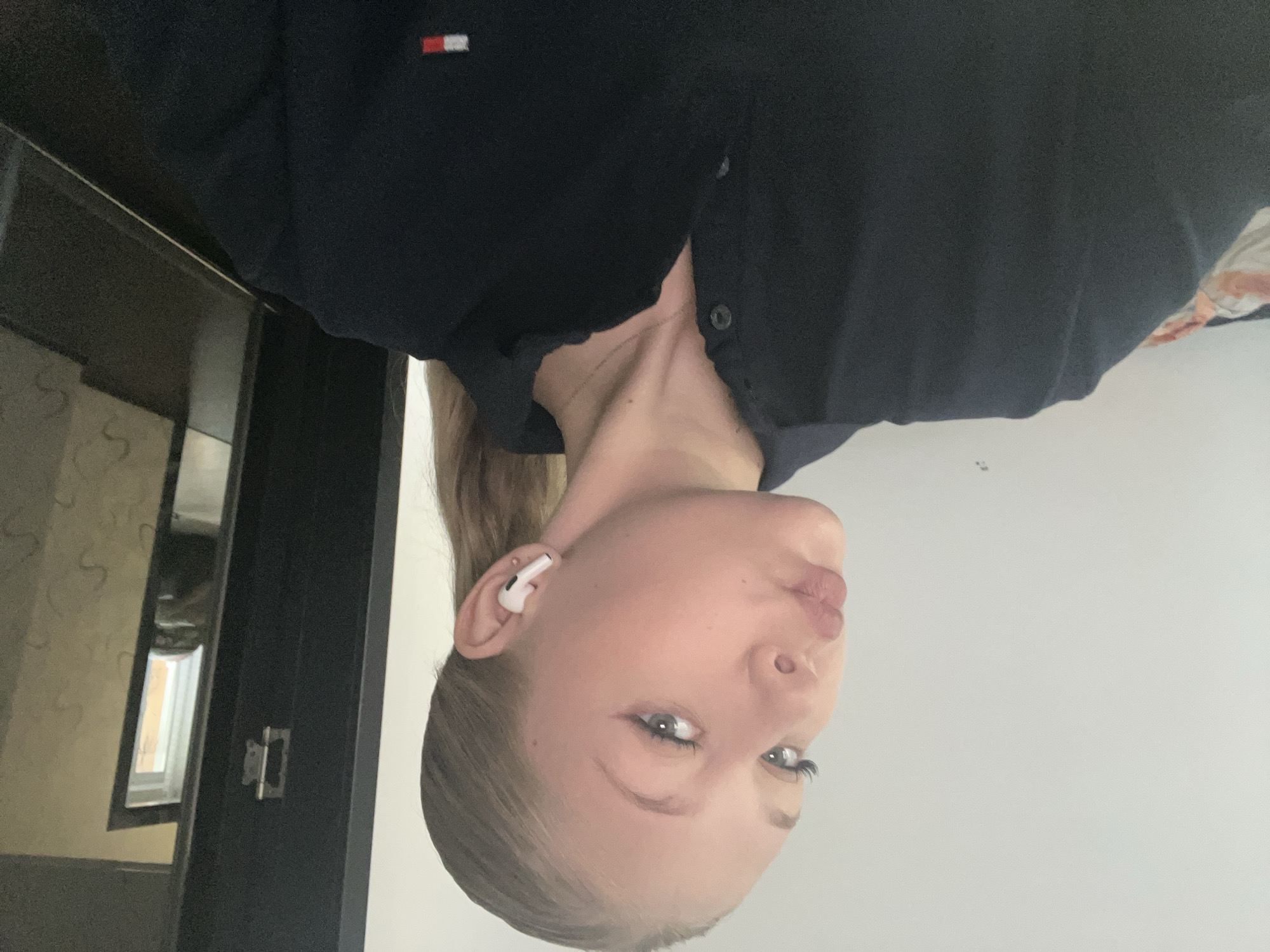 The image shows a young woman with blonde hair pulled back in a ponytail, wearing a dark polo shirt and white wireless earbuds. She is looking at the camera with her lips puckered and her eyes tilted upwards. The background appears to be an indoor setting with a white wall on the left and a dark doorway on the right. The lighting is dim. There is no visible indication of the city of Perm, Russia.