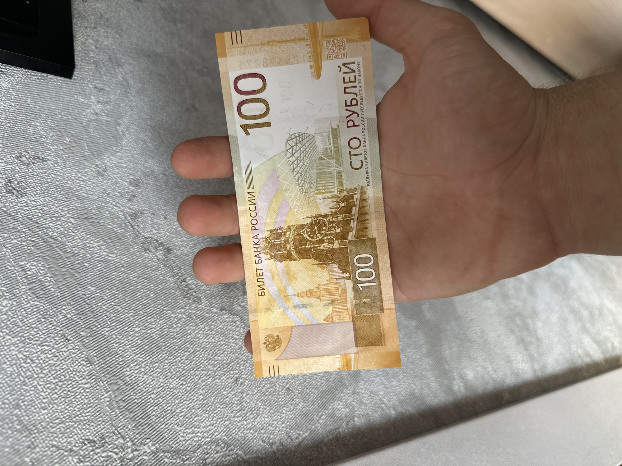 A person's hand is holding a 100 Russian Ruble banknote. The banknote features an image of a clock tower and a modern building. The hand is positioned in front of a textured gray wall. The scene is illuminated by ambient light. There is no discernible location in Birobidzhan, Russia, visible in the image.