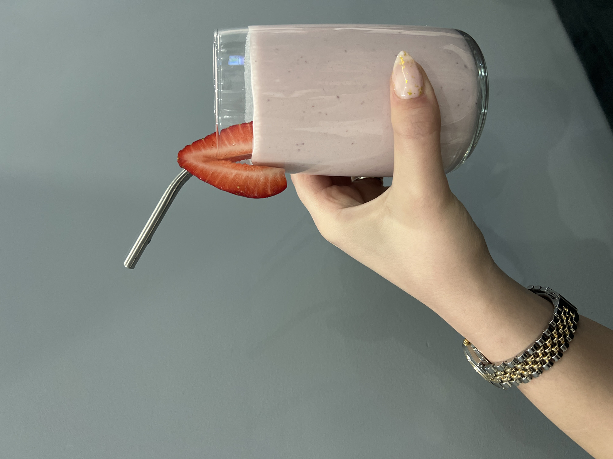 A fair-skinned hand holds a clear glass containing a pink smoothie. A slice of strawberry is partially wedged between the rim of the glass and a stainless steel straw. The hand has a decorative nail with gold accents. A two-tone metal bracelet encircles the wrist. The background is a solid, light gray surface. The location is not discernible from the image, and therefore, it cannot be stated with certainty that this is Yambol, Bulgaria.