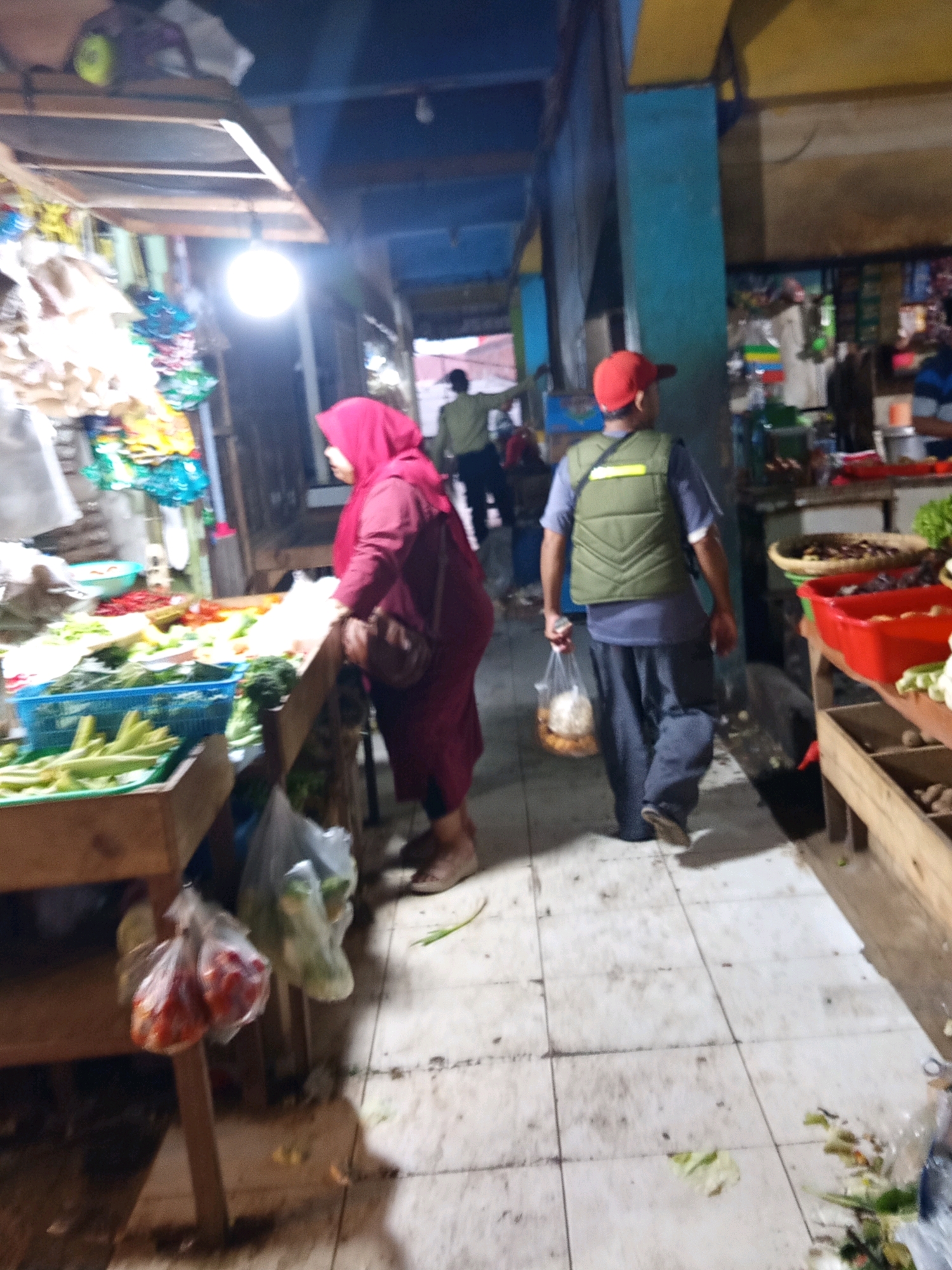 In Karawang, Indonesia, a dimly lit indoor market scene unfolds. A woman wearing a magenta hijab and maroon dress stands by a wooden stall laden with fresh produce, including green vegetables in a blue basket and red chilies. Her right hand rests on the stall. A man in a red cap, a green vest over a gray shirt, and dark pants walks away from the viewer, carrying a clear plastic bag filled with round, light-colored objects. The market floor is tiled with a light-colored grout. Overhead, a bright bulb illuminates the area. Other shoppers and vendors are partially visible in the background. The market appears to be selling a variety of goods, with shelves and displays filled with items.