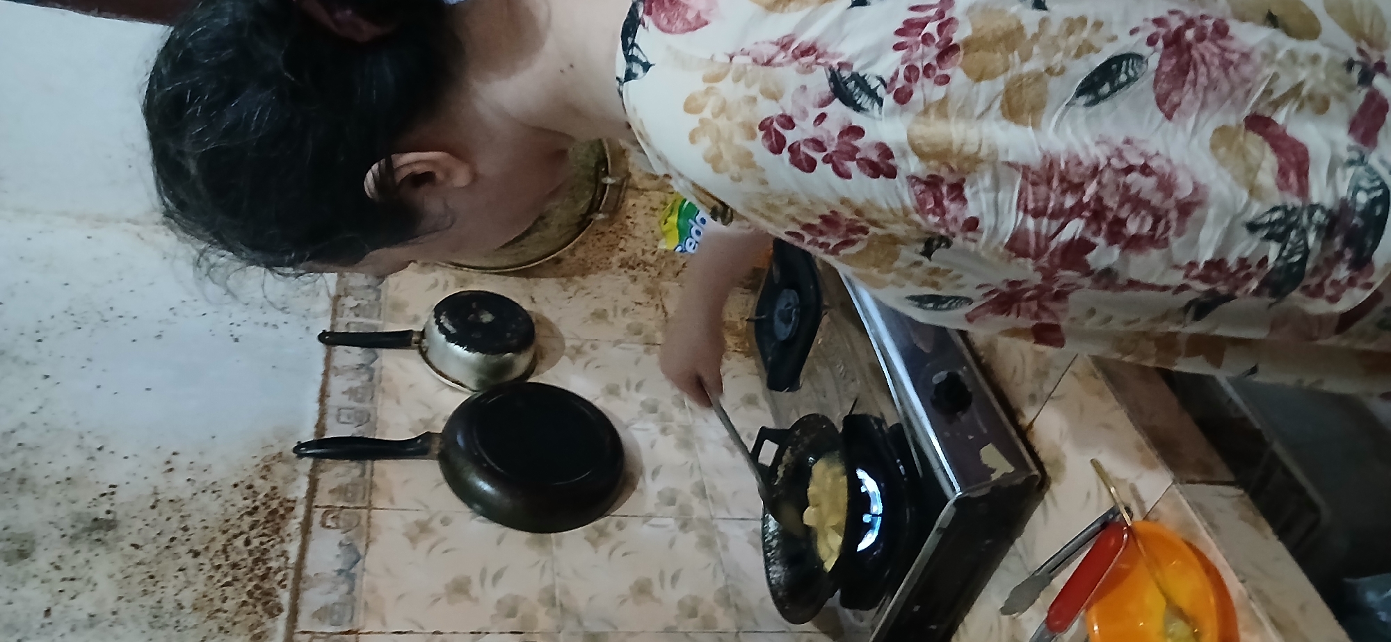 A middle-aged woman, seen from behind, is cooking in what appears to be a modest kitchen in Samarinda, Indonesia. She has dark hair pulled back and is wearing a light-colored blouse with a floral pattern featuring red, yellow, and dark green flowers. Her right hand is holding a spatula, stirring food in a frying pan on a gas stove. The stove is on, with a visible blue flame. Next to the stove, a small orange bowl contains some food and utensils. On the wall behind the stove, two pots are hanging, and the tiled backsplash is visible, showing signs of wear and tear. The wall above the backsplash appears to have water damage or mold. The overall impression is that of a typical home kitchen in a warm climate, likely in a residential area of Samarinda.