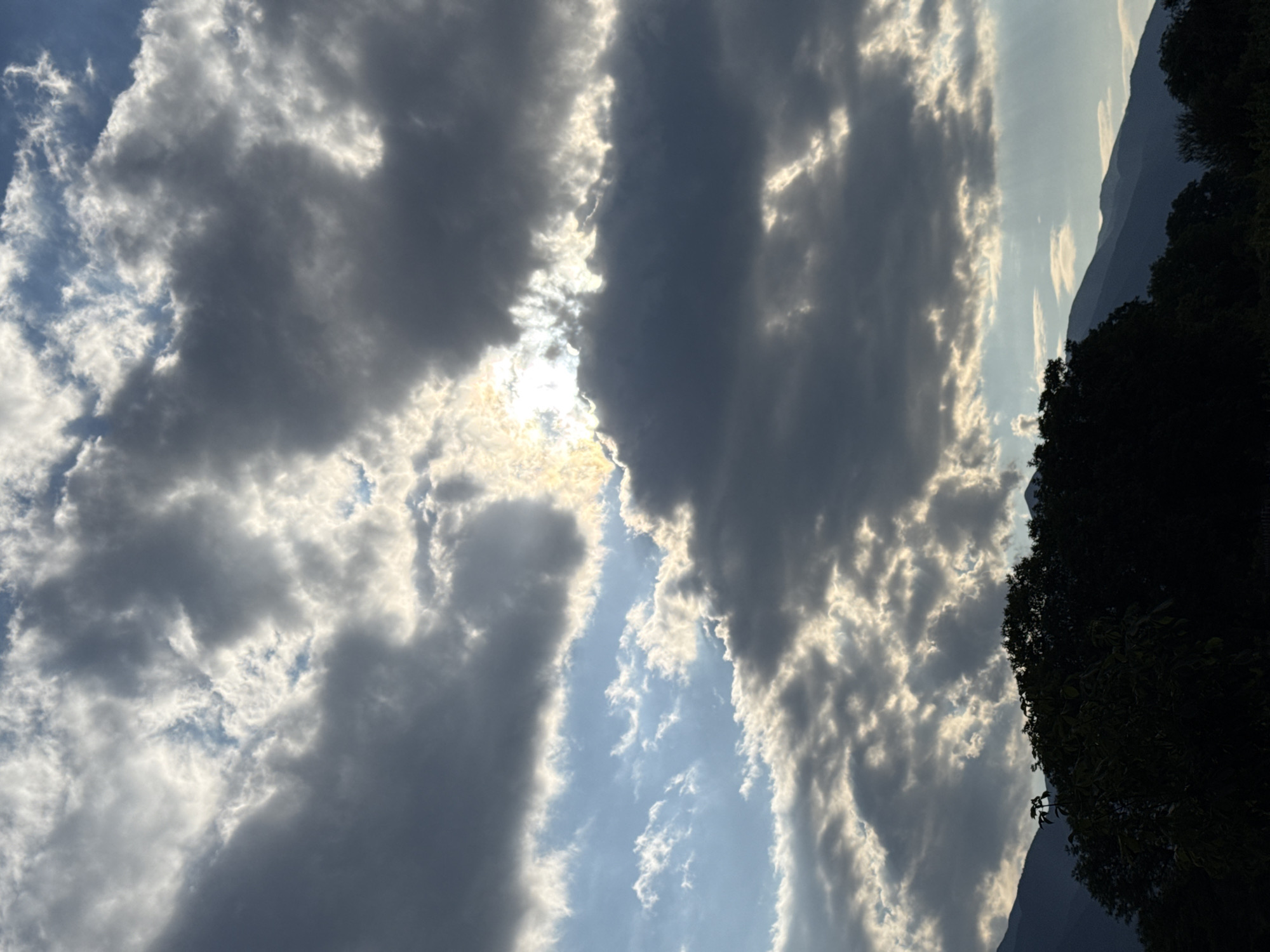 The image depicts a dramatic sky filled with dark, heavy clouds interspersed with patches of bright blue sky and shafts of sunlight. The sun is partially obscured by the clouds, creating a luminous glow. In the foreground, silhouetted against the sky, is a dense line of dark green trees and the dark shapes of distant mountains. The scene captures the moody atmosphere of a potentially stormy or post-storm sky over a mountainous landscape. The location is identified as Bansko, Bulgaria.