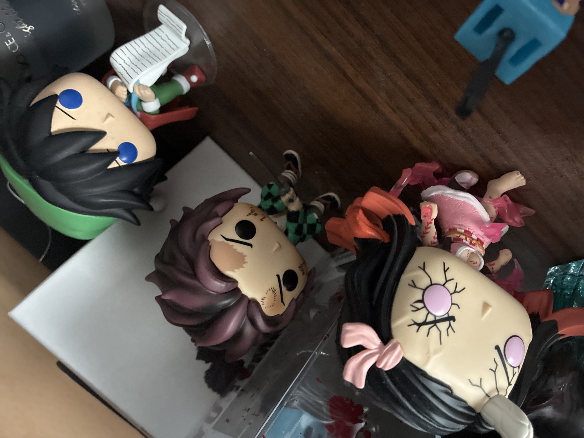 Three anime-style figurines are arranged on a wooden surface. In the foreground, a female figurine with black hair adorned with a pink bow is visible. Her face has distinctive pink circles for eyes and a cracked pattern. To her right, a male figurine with dark hair and a green checkered shirt holds a sword. Behind these two, a third figurine wearing a green hat, red scarf, and green pants is depicted holding a white scroll. The background includes a white box and a dark, reflective object. The figurines are positioned in a close-up, overhead shot. The scene is indoors, on a wooden table. There is no discernible information within the image that suggests a location in Burgas, Bulgaria.