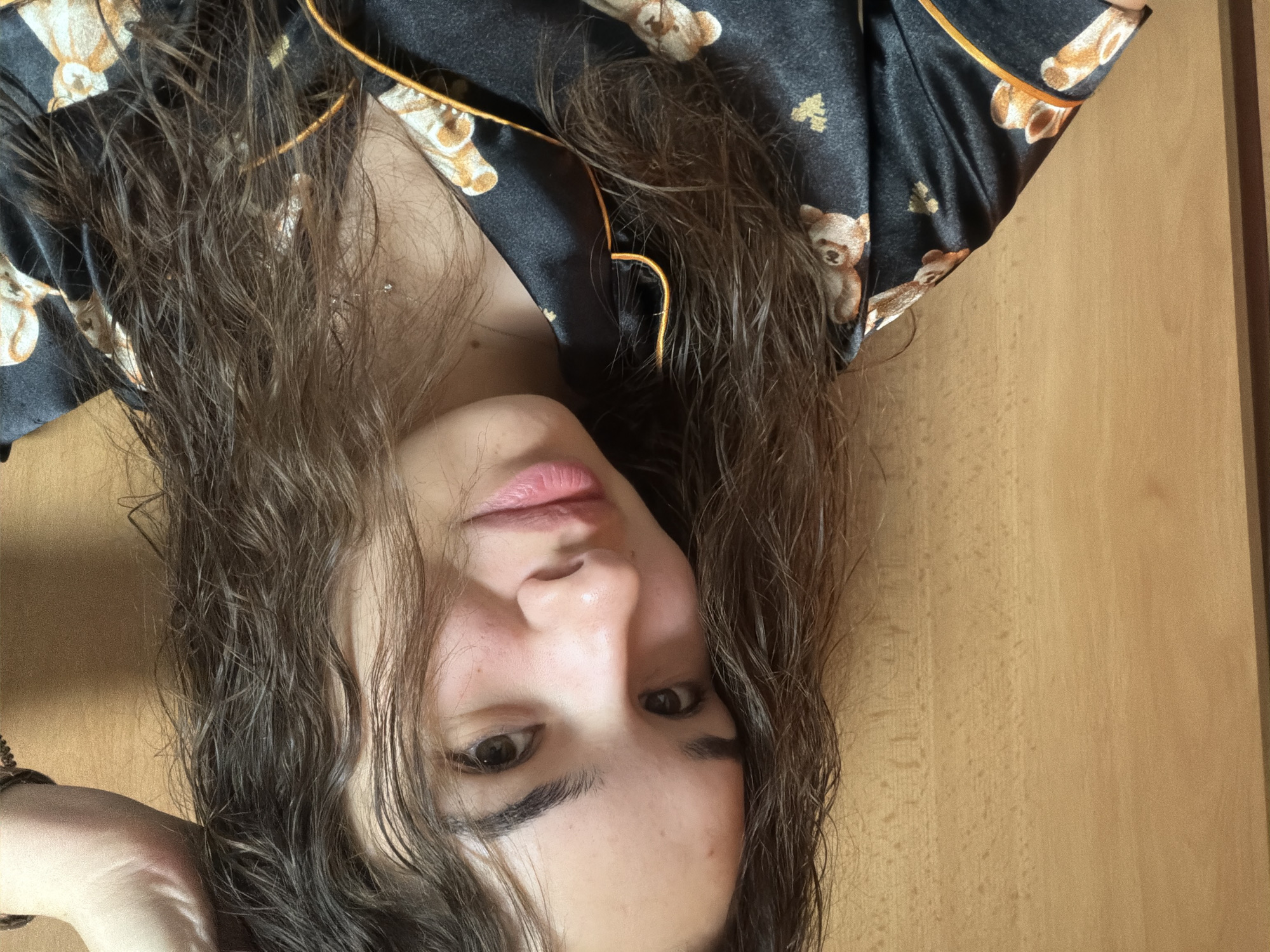 A woman is photographed from a close, upside-down perspective. Her face, including eyes, nose, and lips, is prominently displayed. She has dark hair that is slightly disheveled and resting on her shoulders and chest. She wears a black pajama top with a teddy bear pattern and orange trim. The background appears to be a wooden surface, possibly a door or wall. The photo's location in Sofia, Bulgaria cannot be determined based on this image.