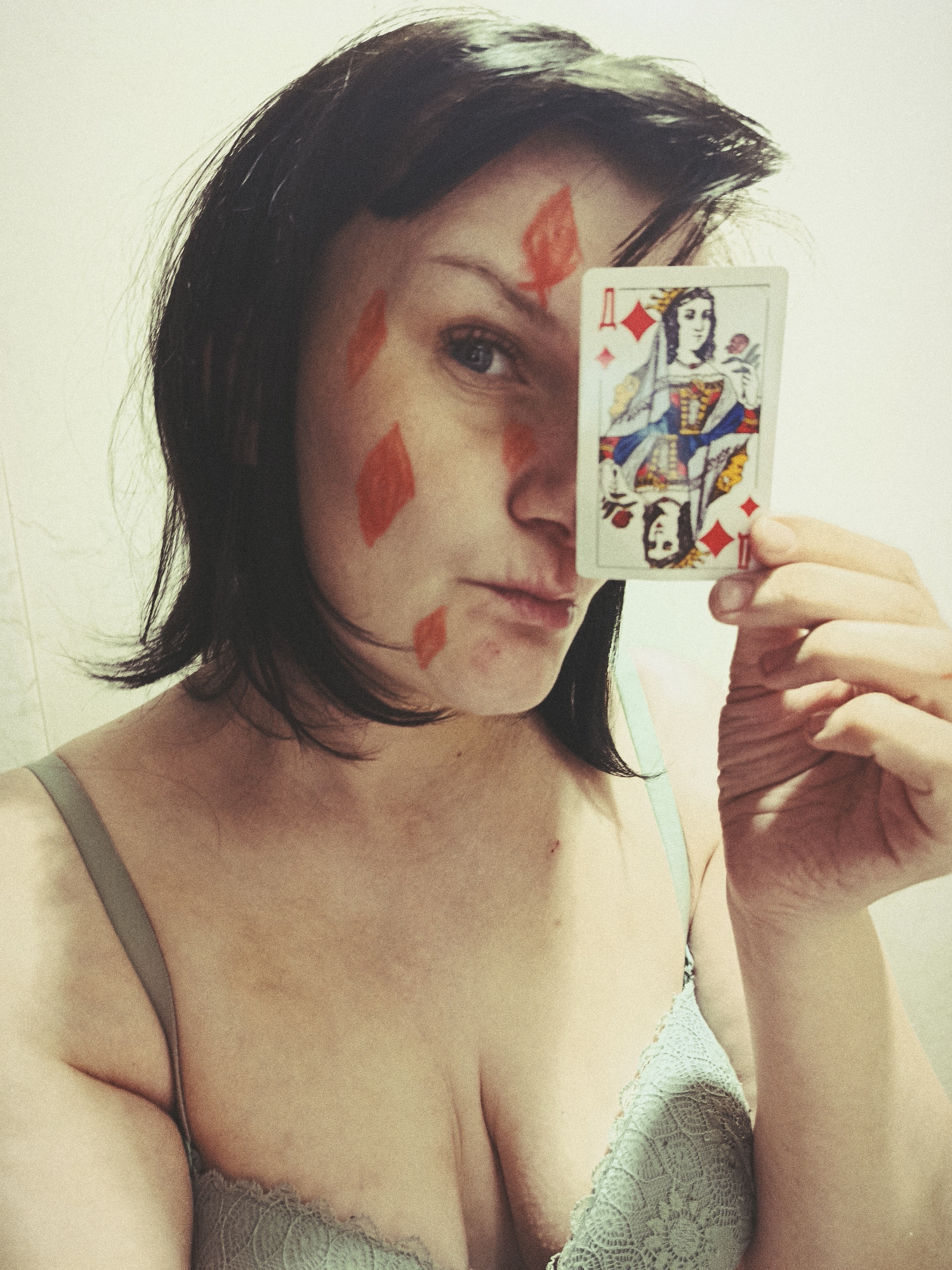 A woman with short, dark hair holds a Queen of Diamonds playing card in front of her face, partially obscuring it. The playing card is positioned vertically, with the image of the queen facing forward. Red diamond shapes are painted on her cheek. She is wearing a light blue, lacy bra. The background is a plain, light-colored wall. There is no evidence that the location is in the city of Фоки, Russia.