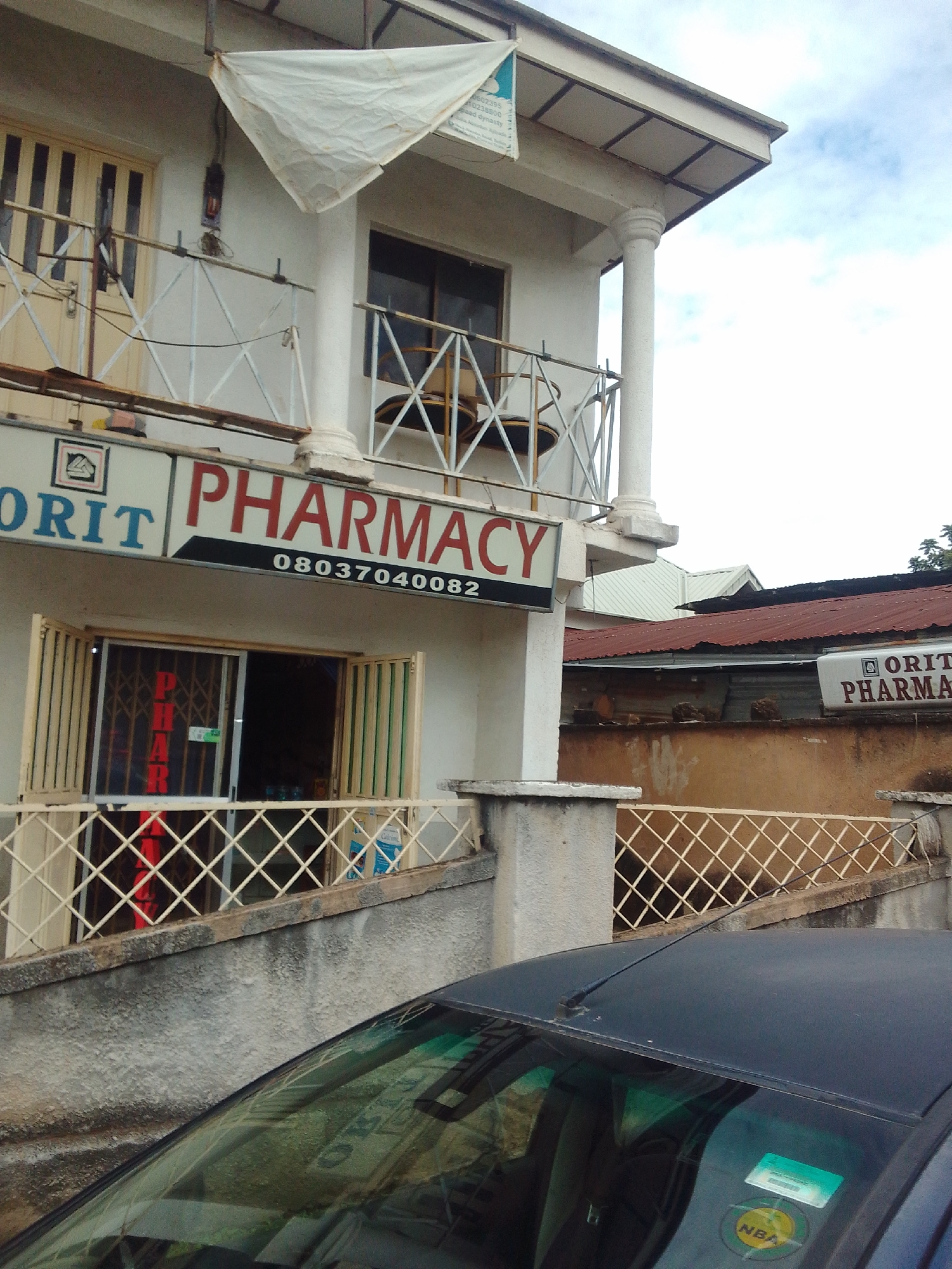 This image captures a street-level view of a two-story commercial building in the city of Jos, Nigeria, featuring an "ORIT PHARMACY." The scene is observed on an overcast day, indicated by the diffused light and light gray sky. The prominent structure is an off-white or cream-colored building. The ground floor houses the "ORIT PHARMACY," marked by a large horizontal sign with "ORIT" in blue letters and "PHARMACY" in red letters, set against a white background with a black border. Below "PHARMACY," a black strip displays the phone number "08037040082." The pharmacy entrance is secured by a partially open, light-colored accordion-style metal security gate with a diamond pattern. Behind this gate, through the open doorway, vertical red letters spelling "PHAR" are faintly visible, likely part of an interior sign. To the right of the entrance, display windows reveal various packaged products. A low, off-white metal railing with a diamond pattern runs along the front of the ground floor. Below this railing is a textured, weathered gray concrete wall. The upper floor of the building features a balcony area supported by a prominent white, fluted column with a decorative capital. To the left, a set of closed, light-colored double doors with dark vertical panels is visible, fronted by a matching off-white metal railing. To the right of the column, a dark-paned window is similarly fronted by a metal railing. A small, dark, possibly circular object or fixture is visible behind the railing within the window frame. From the roof overhang above, a triangular piece of white fabric is suspended, partially obscuring a vertical banner with faint text and numbers, including "0602395" and "023800." In the immediate foreground, the hood and windshield of a dark-colored (likely black or dark blue) car are visible. The windshield has a green tint at the top and reflects parts of the building facade. Two stickers are affixed to the passenger side of the windshield: a rectangular green one at the top and a circular yellow one with "NBA" printed on it at the bottom right. A black radio antenna extends from the car's roof near the windshield. In the background, beyond the main building, several rooftops are visible, including a rusty-red corrugated metal roof and a lighter gray corrugated metal roof. Green foliage from trees is visible on the far right. Another smaller, white sign with "ORIT PHARMA" in brown letters is partially visible on a building