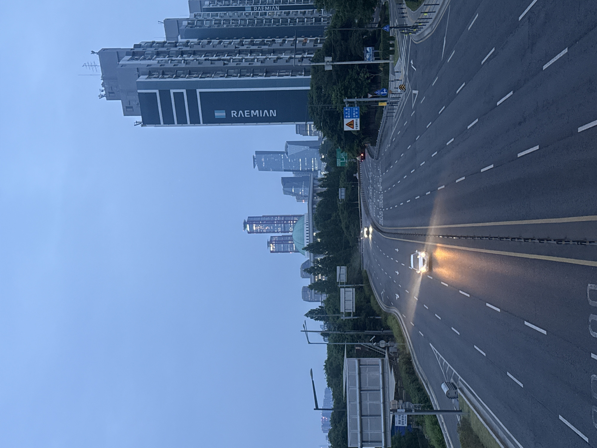 The image depicts a highway scene in Seoul, South Korea. It is an elevated view, with multiple lanes of traffic visible. A car with headlights on is approaching. On the left side of the road, there is a modern building with "RAEMIAN" written on it. In the background, the skyline of the city is visible. The sky is a pale blue, indicating either dawn or dusk. There are road signs and streetlights present. The road is marked with white and yellow lines. Greenery is visible along the roadside.