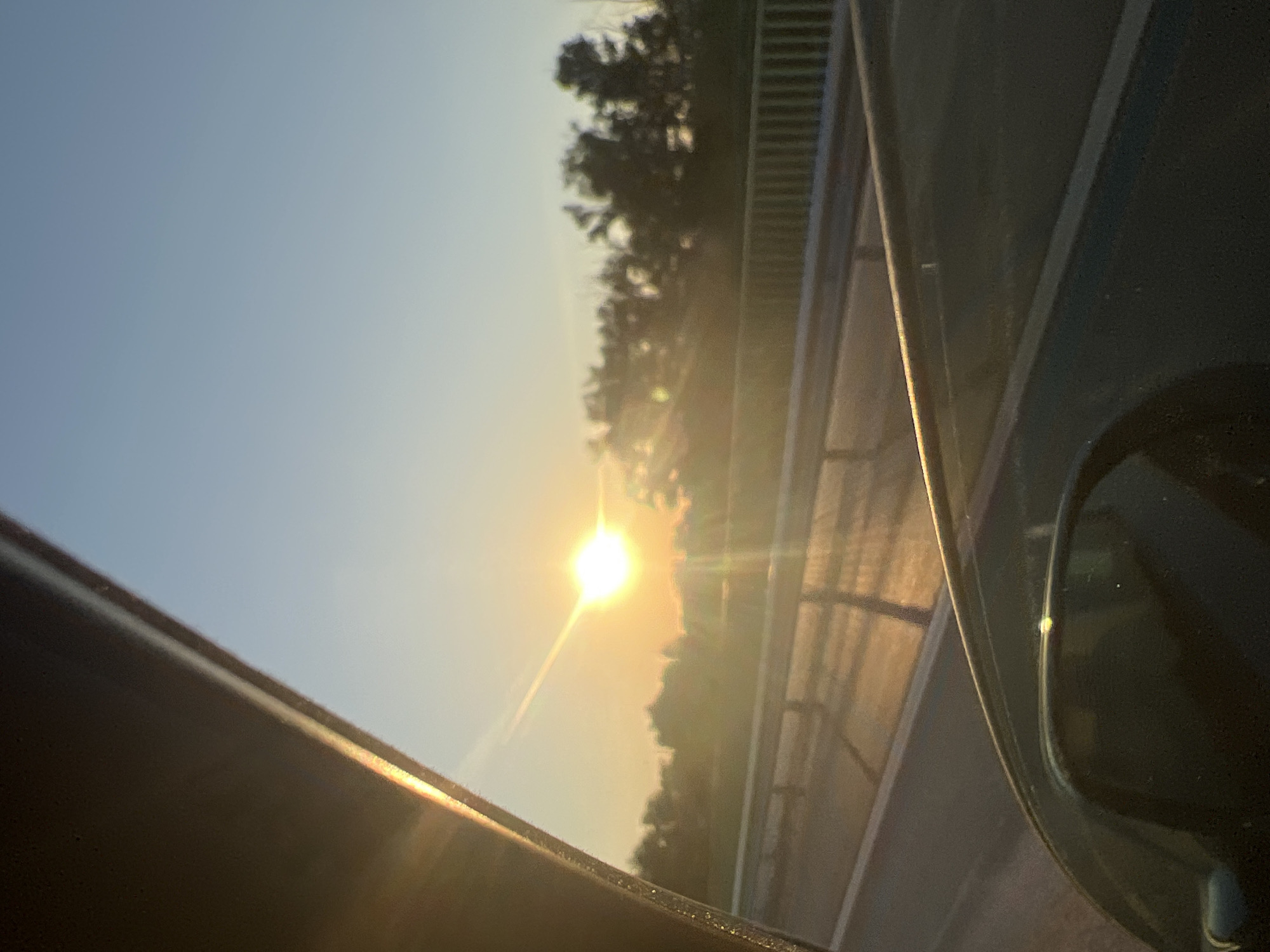 The image shows a sunset viewed from inside a vehicle, likely from the passenger seat, looking out the side window. The sun, a bright orb with visible lens flare, is low on the horizon, casting a warm, golden light across the scene. The sky is a clear, soft blue, transitioning to a lighter hue near the horizon. In the middle ground, a treeline is silhouetted against the bright sky, with some foliage visible. A guardrail runs parallel to a road, suggesting the vehicle is on a highway or major road. The foreground is dominated by the interior of the car, with the car's window frame and a portion of the side mirror visible. The overall impression is one of a peaceful, scenic drive during golden hour. The location, Noven'kii, Russia, is not visually discernible from the image itself.