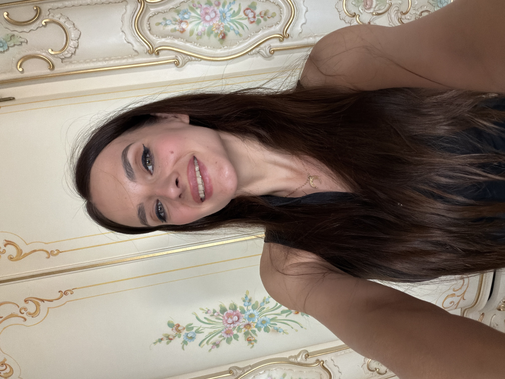 A woman with long, dark brown hair is taking a selfie. She is smiling with her mouth slightly open, revealing her teeth. She is wearing a black top and a gold necklace with a crescent moon pendant. The background appears to be a wall with ornate floral and gold detailing, possibly a closet or a decorative panel. The lighting suggests it is daytime and the location is indoors.