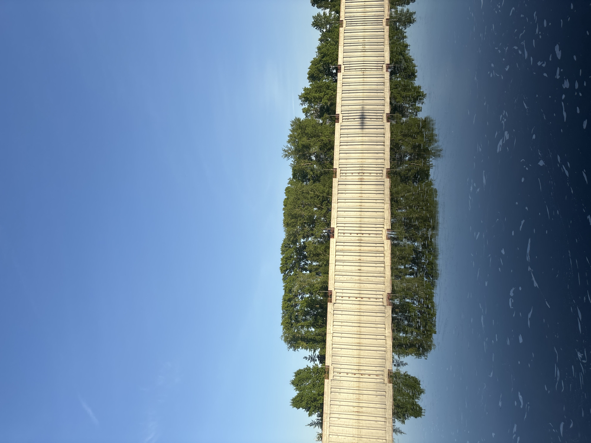 The image shows a pier in Chaikovsky, Russia, stretching into a body of water. The pier is bordered by trees on each side. The left side of the image is a clear blue sky, while the right side shows the water with reflections of the pier and trees. The pier is constructed of light-colored planks, and its shadow is visible on its surface.